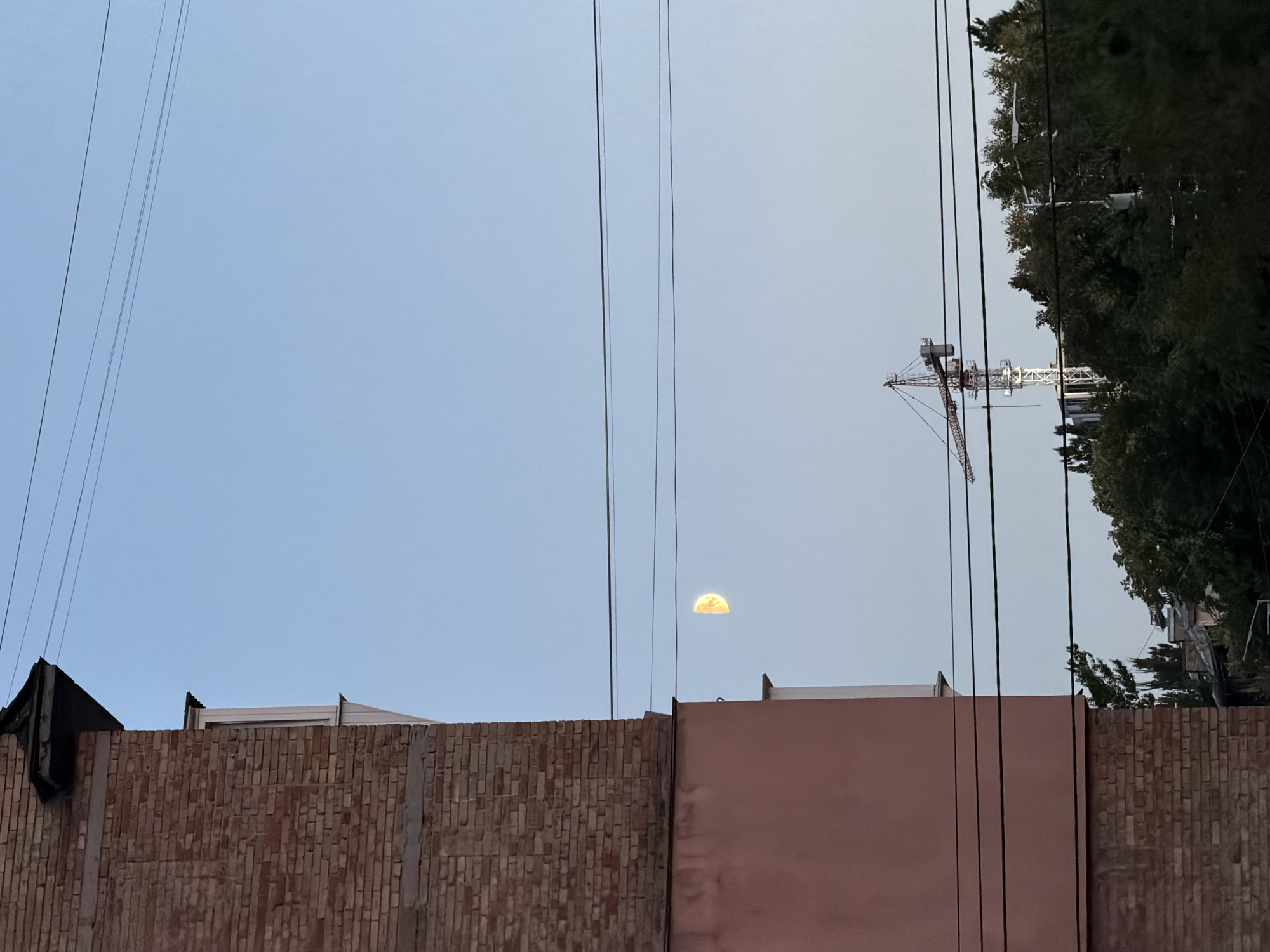 A half-moon is visible in a pale blue sky, slightly to the left of the center of the frame. In the foreground, on the left, is the side of a building constructed of red brick with a flat roof. Adjacent to this is a section of the building painted a muted pink. Three black wires stretch horizontally across the sky. In the middle ground, above a line of dark green trees, a construction crane with a red boom is visible. In the background, other buildings and trees can be seen. The scene appears to be an urban environment. The perspective suggests the photo was taken from a window or balcony.