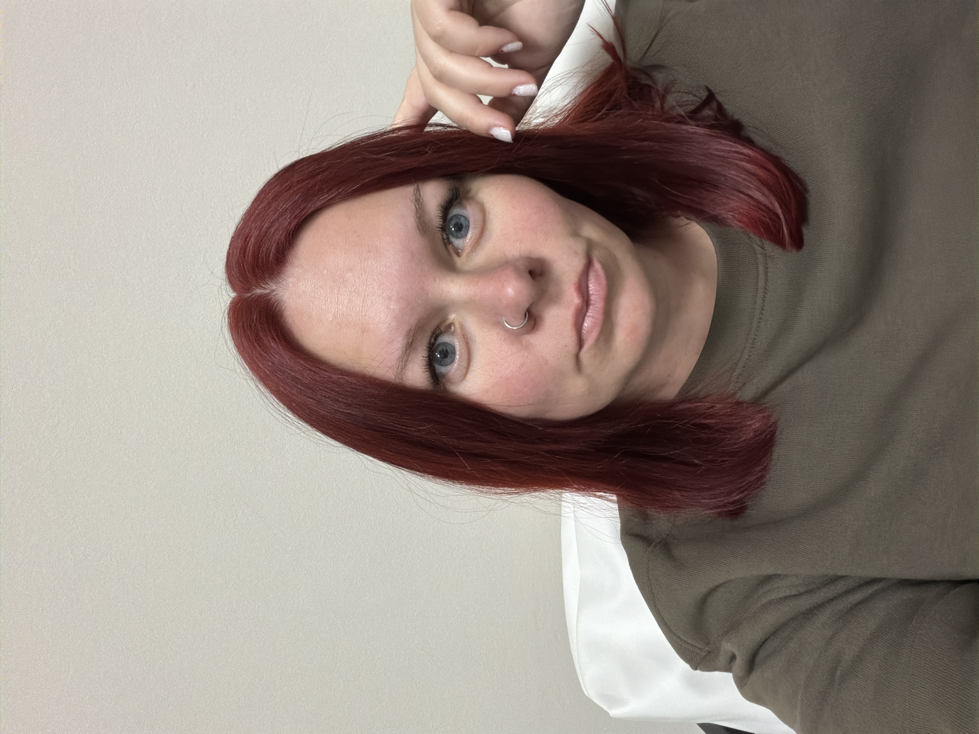 An adult female is depicted in a close-up, chest-up shot, presumably located indoors in the city of Chaikovsky, Russia. The subject has shoulder-length, dark red hair, parted centrally, and blue eyes. A silver-colored hoop nose ring is present on her left nostril. She is wearing an olive-green, long-sleeved crew-neck top. The subject faces forward, looking directly into the camera with a neutral expression and slightly parted lips. Her left hand is positioned with fingers resting near the right side of her head, above the ear; her fingernails are painted with a light, possibly glittery, polish. The background consists of a plain, light-colored wall with a subtle texture. A white object, possibly a chair back or cushion, is partially visible behind her left shoulder. Lighting is even.