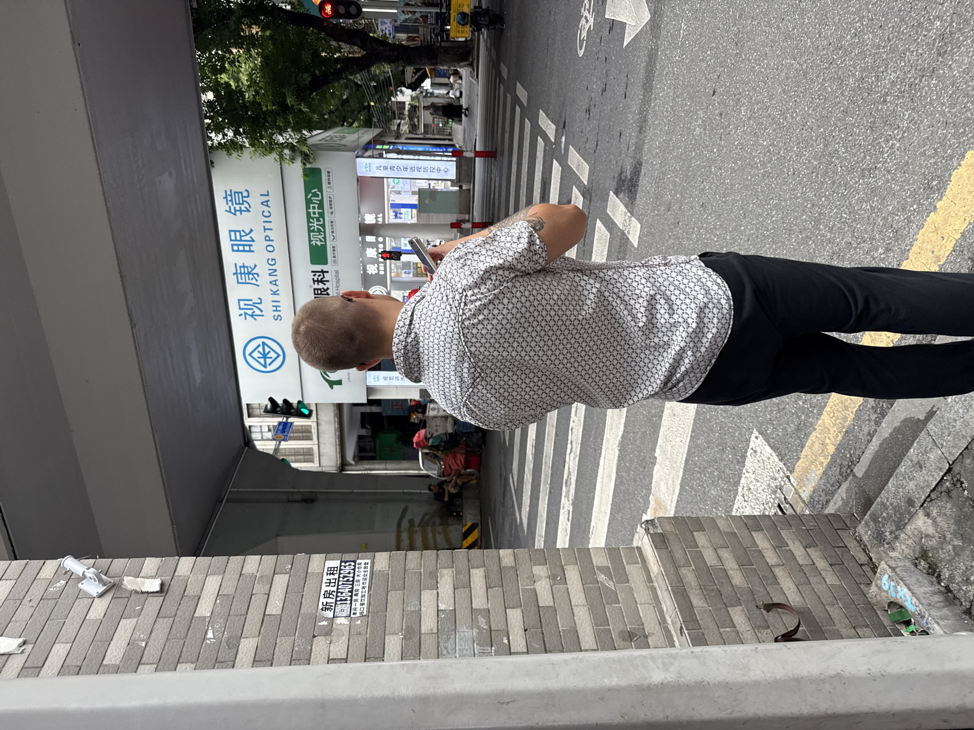The image shows a street scene in Yuexiu District, China, under an overpass. In the foreground, a man is standing facing away from the camera, looking at his mobile phone. He is wearing a patterned short-sleeved shirt and black pants. Behind him, a pedestrian crossing is visible, with a traffic light showing green in the distance. A building with signage that reads "SHI KANG OPTICAL" is also present. Further back, several people appear to be sitting or standing near what looks like a makeshift stall or collection of belongings. The overall environment suggests a busy urban area.