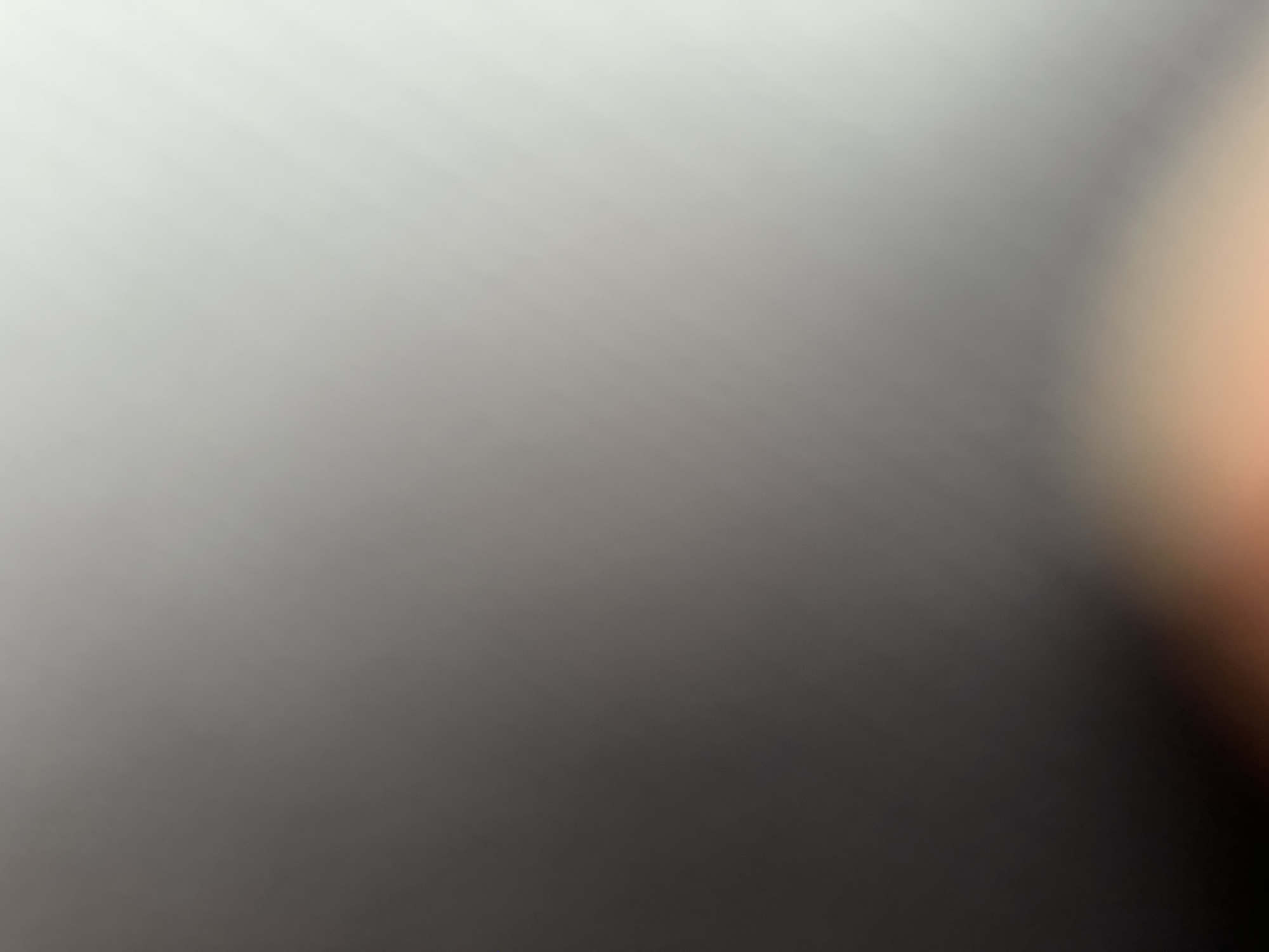 This is a blurred, abstract image with a gradient of colors ranging from light gray to dark gray, with a soft orange glow on the right side. There are no discernible subjects, objects, or a clear scene or setting. No activity or event is taking place. There are no notable details such as time of day, weather, location cues, or emotions. There is no visible text. The image appears to be a close-up of a surface or a lens flare, with no specific context provided.