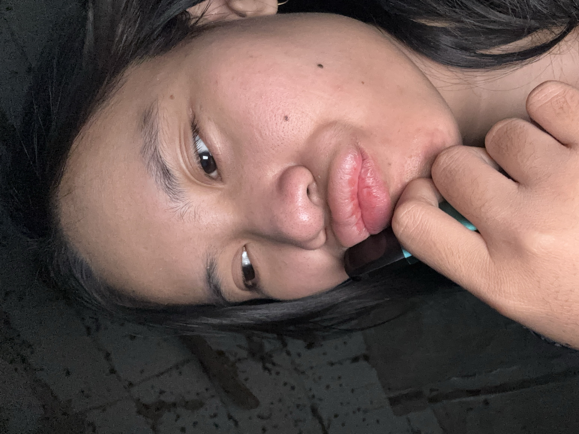 A close-up shot shows a young woman of East Asian descent with dark hair, a mole on her right cheek, and full lips puckered as she holds a black and teal object to her mouth. She is looking off to the left, with her right hand supporting her chin. The background is dark and textured, resembling a concrete wall. The lighting is dim, creating shadows and highlighting her facial features. The image appears to be a selfie, taken in an indoor environment.