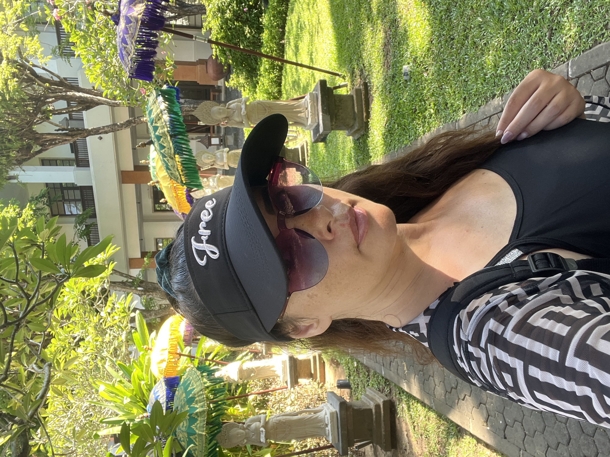 A selfie features a woman with brown hair, wearing a black visor with the word "Free" embroidered in white, and pink-tinted sunglasses. She is also wearing a black tank top and a black and white patterned mesh top over it, with a backpack strap visible. The background shows a pathway lined with stone statues and ornate umbrellas, with lush greenery and a building in the distance. The setting appears to be a garden or courtyard, possibly in Kuta Selatan, Indonesia, given the style of the statues and umbrellas, which are characteristic of Balinese decor. The lighting suggests it is daytime, with sunlight filtering through the trees.