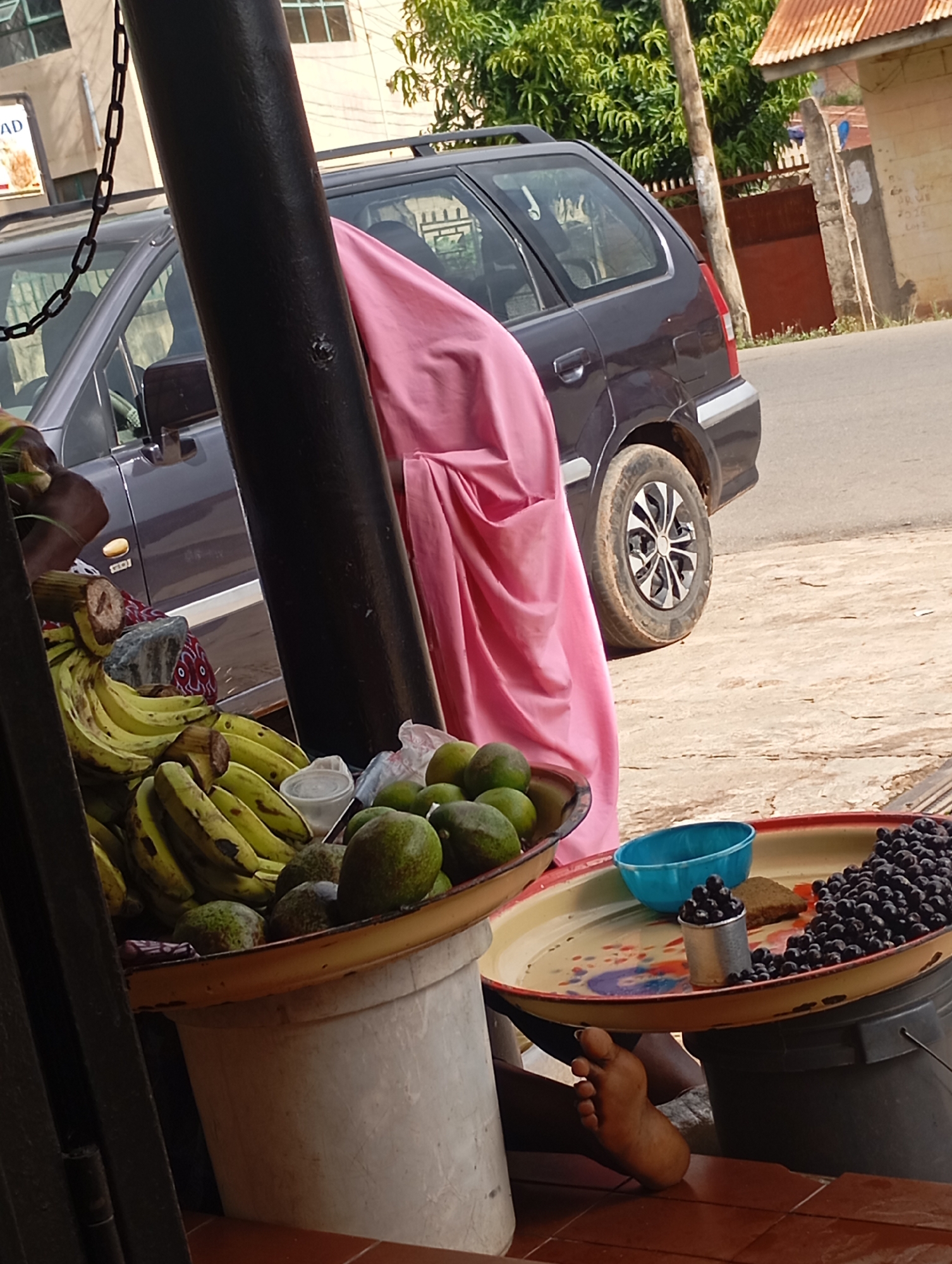 A person's bare foot is visible on the right side of the image, placed on a tiled surface. In the center, a large metal tray contains a pile of dark berries and a small blue bowl. The tray is positioned over a black bucket. To the left of the tray, a large, light-colored container holds bananas and several green avocados. A dark, thick pillar is prominent in the foreground on the left. Behind the pillar, a dark-colored car is parked. The scene suggests a roadside market stall. This image likely depicts a scene in Jos, Nigeria, given the context of typical street vending and the vehicle type.