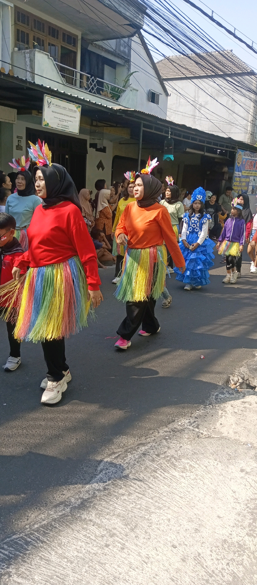 This image captures a street scene in Malang, Indonesia, likely during a festive event or parade. The foreground is dominated by several people dressed in vibrant costumes. Two women in the center wear black hijabs and brightly colored, layered skirts made of fringed material in rainbow hues. The woman on the left wears a red long-sleeved shirt, while the woman to her right wears an orange long-sleeved shirt. Both have feathered headpieces. Behind them, a young person in a blue, ruffled dress and a matching headpiece is visible, as is another person in a purple jacket and a similar colorful skirt. The background shows buildings with balconies and tiled roofs, suggesting a residential or commercial area. Overhead, a tangle of electrical wires crisscrosses the sky, a common sight in Indonesian cities. The street surface is asphalt, with some areas of concrete and shadows cast by the sun. The overall atmosphere suggests a lively, outdoor gathering.