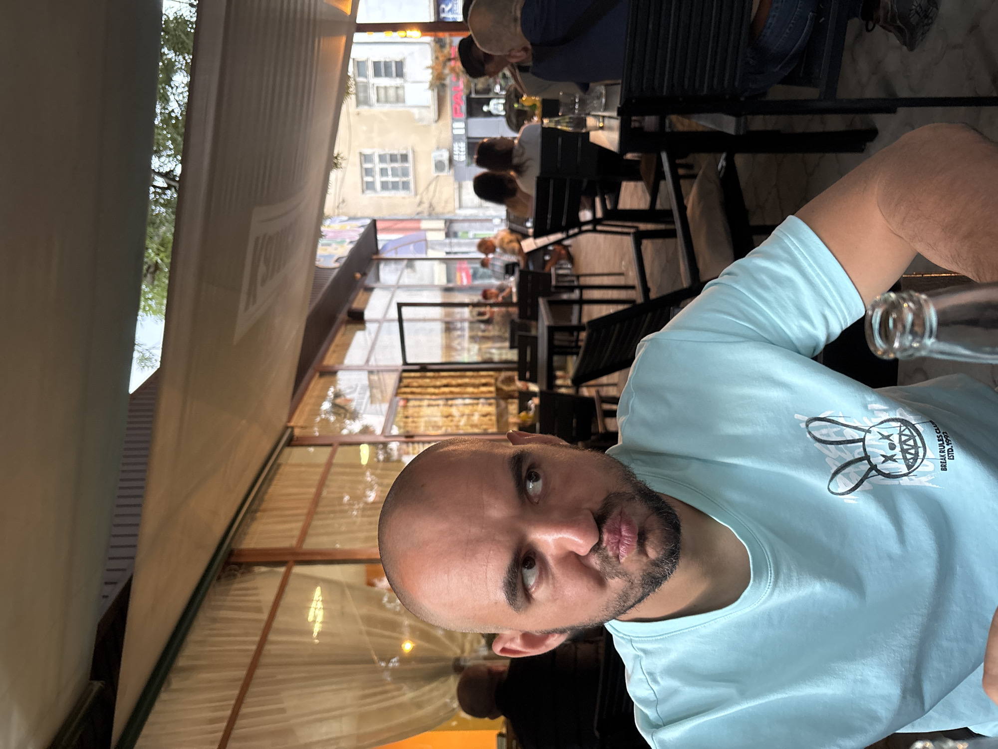 An outdoor cafe scene in Karlovo, Bulgaria, is depicted. In the foreground, a bald adult male with a short beard is seated, wearing a light blue t-shirt featuring a stylized bunny graphic with crossed eyes and sharp teeth, and the text "BREAK RULES CLUB ESTD. 1992." He is looking to his left (viewer's right) with pursed lips, exhibiting a distinct facial expression. A clear glass bottle is partially visible on the table in the lower left.
The midground shows an active cafe environment with multiple patrons, most of whom are blurred, seated at dark tables with matching chairs. A person wearing a dark shirt is partially visible to the primary subject's right (viewer's left). Behind the seated individuals, a building with large windows and light-colored interior curtains suggests an indoor section of the establishment or an adjacent building.
The background reveals an urban street with older buildings. One building facade displays fragmented commercial signage including "JEANS WEAR" and "PAL...". Graffiti or street art is visible on a wall in the upper left portion of the background. The outdoor seating area is covered by a light-colored, possibly retractable, awning. Above the awning, a narrow strip of trees and clear sky is visible.
