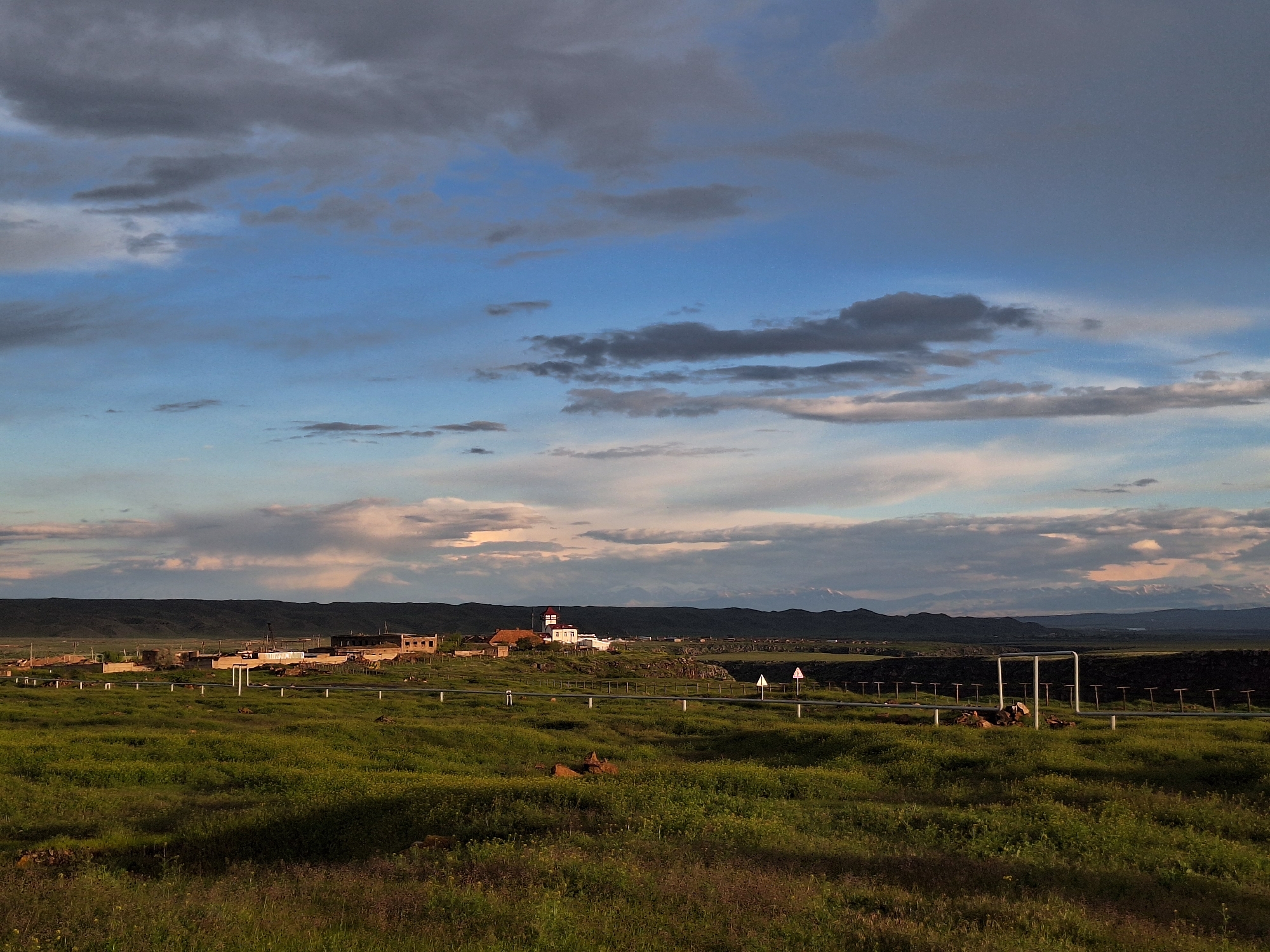 The image shows a landscape in or near the city of Anipemza, Armenia. In the foreground is a field of green grass with some wildflowers. A slightly elevated horizon line runs horizontally across the middle of the image, supporting structures that include houses and a church with a red roof. A raised pipeline and warning signs bisect the field, running roughly parallel to the horizon. In the background are mountains, with what appear to be snow-capped peaks in the distance. The sky is filled with blue and grey clouds with patches of light.