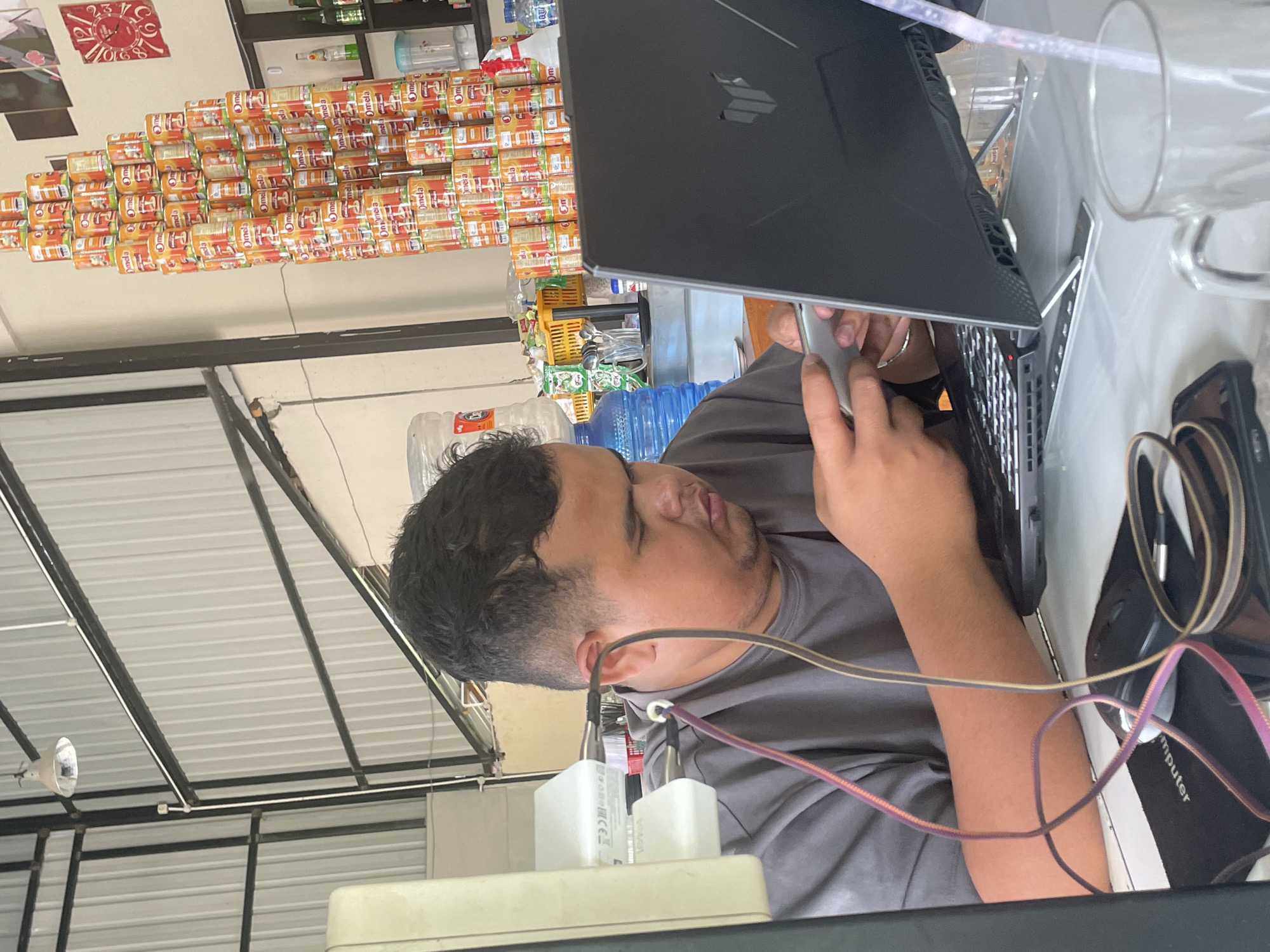 In Pasar Paloh, Indonesia, a young man is seated at a table, engrossed in his phone and a laptop. He is wearing a grey t-shirt and has short, dark hair. The table is cluttered with electronic devices, including the laptop, a phone, and a computer mouse. Wires are connected to his earbuds, which are plugged into both his phone and a wall adapter. Behind him, a tall stack of canned goods, labeled "Omela," dominates the background, reaching towards the ceiling. The environment appears to be an indoor space with industrial-style metal roofing and lighting fixtures. A large blue water dispenser is also visible, along with various shelves containing bottles and other items.