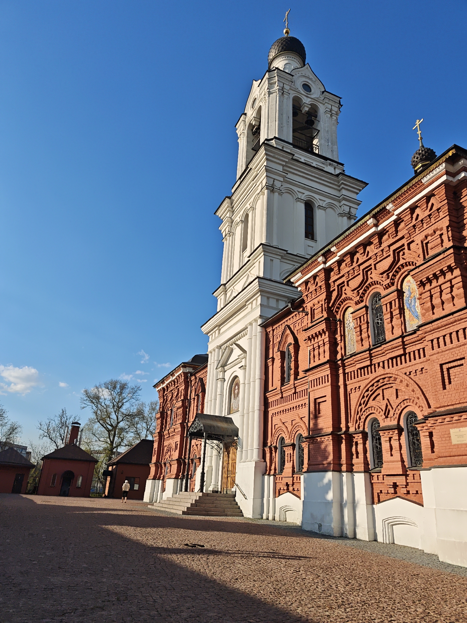 The image depicts the Bogoyavlensky Cathedral in Noginsk, Russia, under a clear blue sky with a few scattered clouds. The cathedral complex is composed of a red brick building with intricate architectural details and a white bell tower. A single figure can be seen walking on the cobblestone courtyard. There are also three small red brick buildings with dark roofs. The sun casts long shadows across the scene.