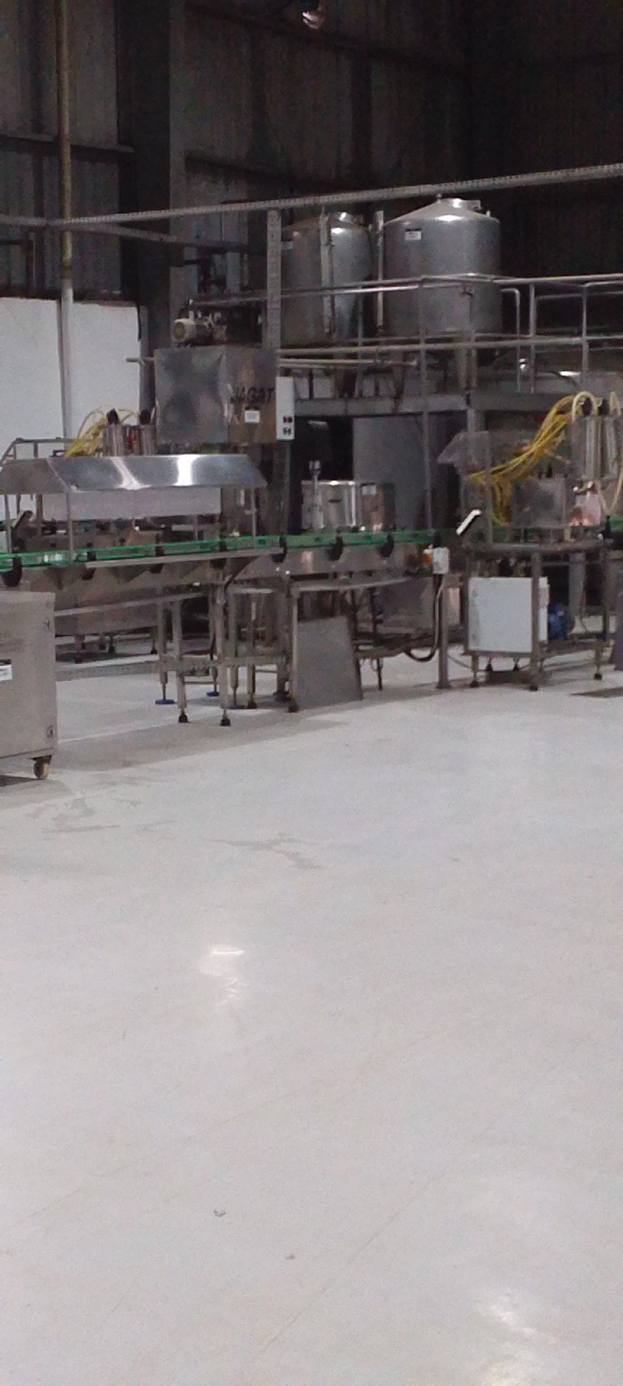The image shows an industrial processing facility with a conveyor belt system running horizontally across the middle of the frame. The system appears to be made of stainless steel and features various components, including tanks, containers, and machinery. Overhead, a metal structure supports more tanks and equipment. The environment is a large, open warehouse with a light-colored floor. The location is not specified, and the city of Unknown, Unknown, is fictional.