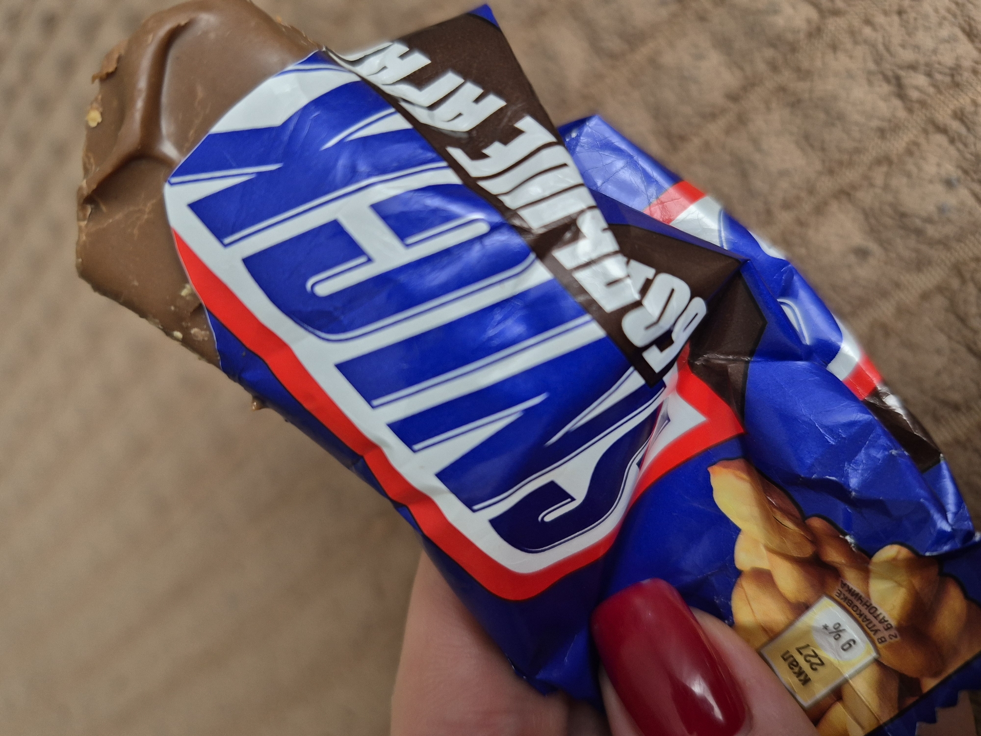 A partially unwrapped Snickers bar is held in a hand with a red fingernail. The wrapper is blue, white, and red and features the word "SNICKERS." Nutritional information is visible on the lower right of the wrapper, written in Russian, suggesting the location may be in Russia, possibly near Tobolsk. The background is a light brown fabric.