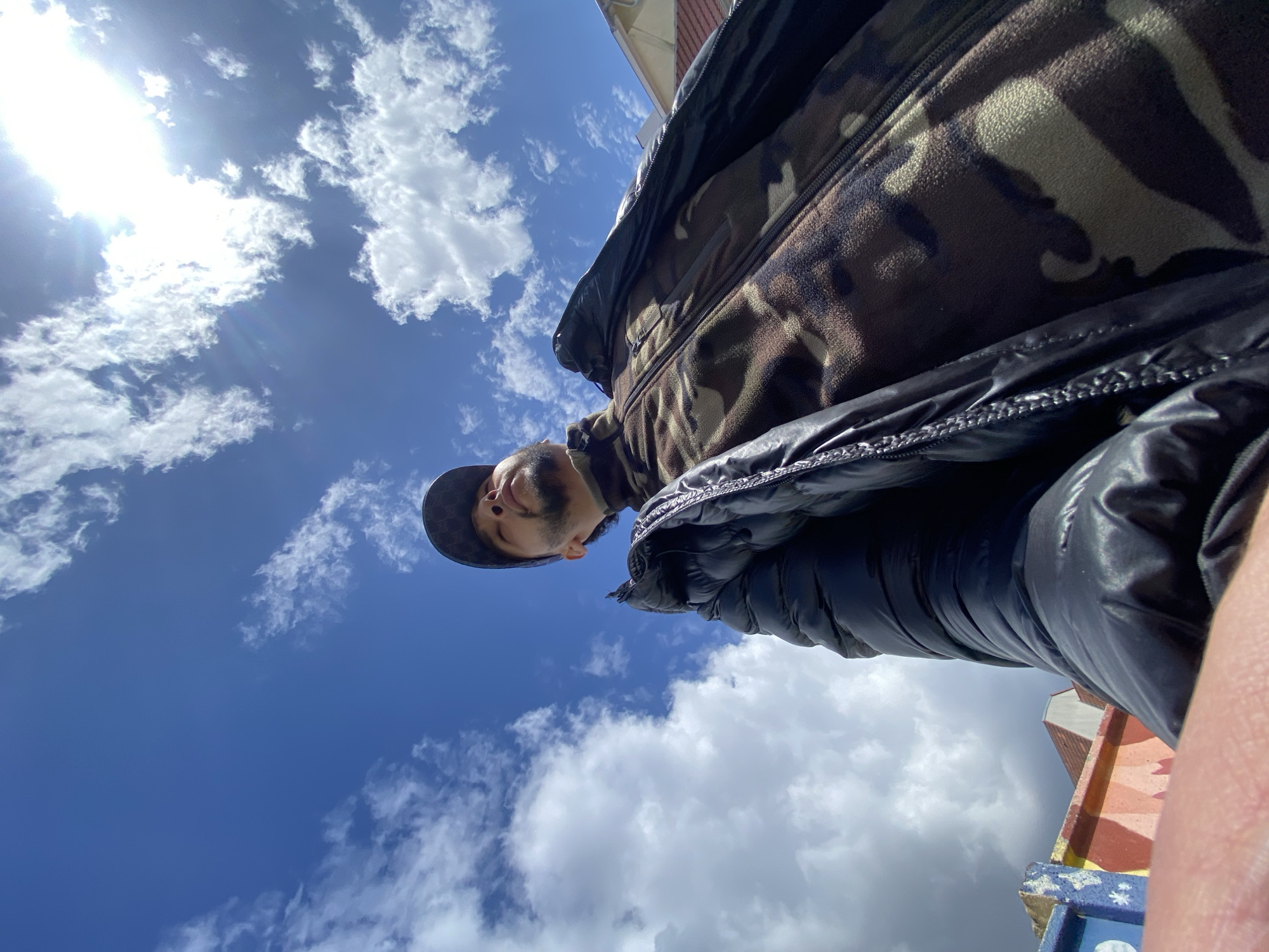 A low-angle shot looking up at a person wearing a cap, camouflage fleece jacket, and a puffy black down jacket in Bischheim, France. The person is angled towards the upper left, with blue sky and white clouds filling the background. Part of a building with a red tile roof and colorful mural is visible in the lower right corner. The sun is shining brightly in the upper left.