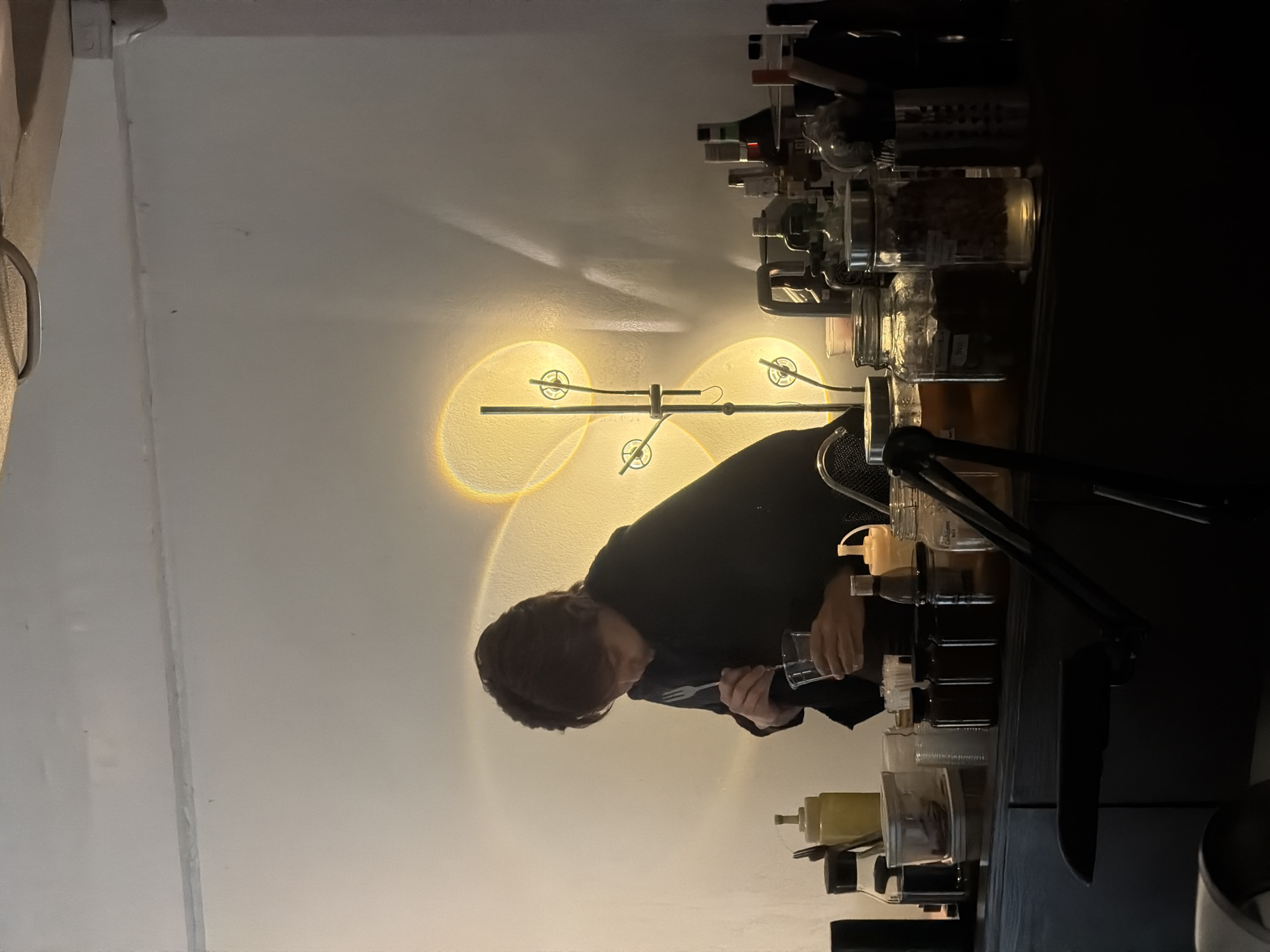 The image depicts a dimly lit interior, likely a bar or cafe, with a person dressed in black working behind a counter. The person is holding a clear plastic cup and appears to be mixing something with a utensil. Behind them, a wall is illuminated by a decorative lamp that casts a soft, warm glow, creating circular patterns of light. The counter is lined with various containers, including glass jars filled with what appear to be dried ingredients, bottles, and dispensers. To the right, several bottles, some with dark liquid, are arranged on the counter. The overall ambiance is intimate and possibly nocturnal, suggested by the low lighting and the presence of bottles that might contain alcoholic beverages. The scene is devoid of any specific identifying landmarks that would definitively place it within Chiang Mai City Municipality, Thailand, but the style of the bar and the arrangement of goods could be consistent with establishments found in such a location.