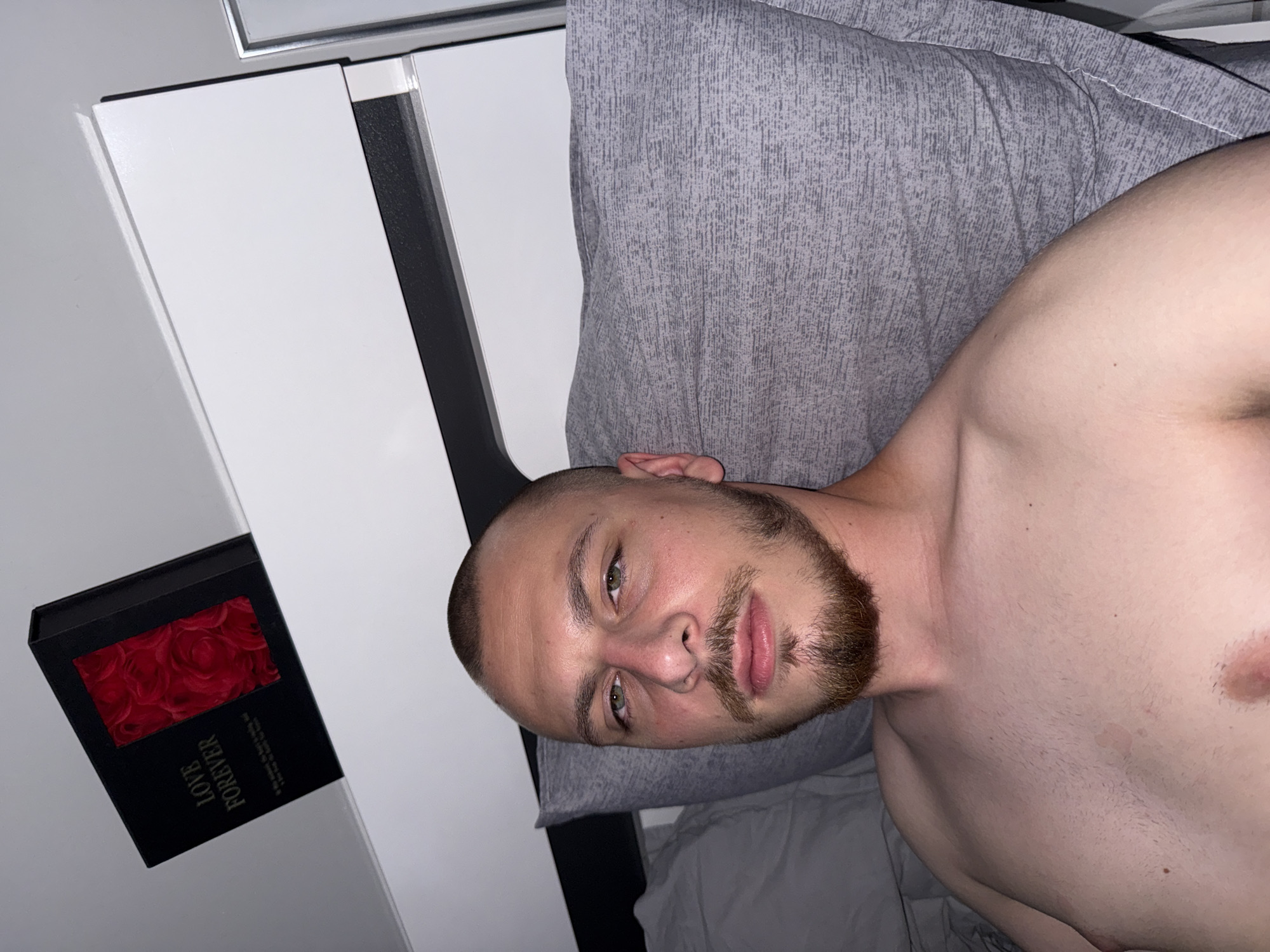 The image shows a close-up, selfie-style shot of a man lying in bed. He has a shaved head, green eyes, and a goatee. He is shirtless, and his chest and shoulders are visible. He is lying on a grey pillow, and the background appears to be a bedroom with a white headboard. On the headboard, there is a black box with the words "LOVE FOREVER" and an image of red roses. The image is not in Stara Zagora, Bulgaria.