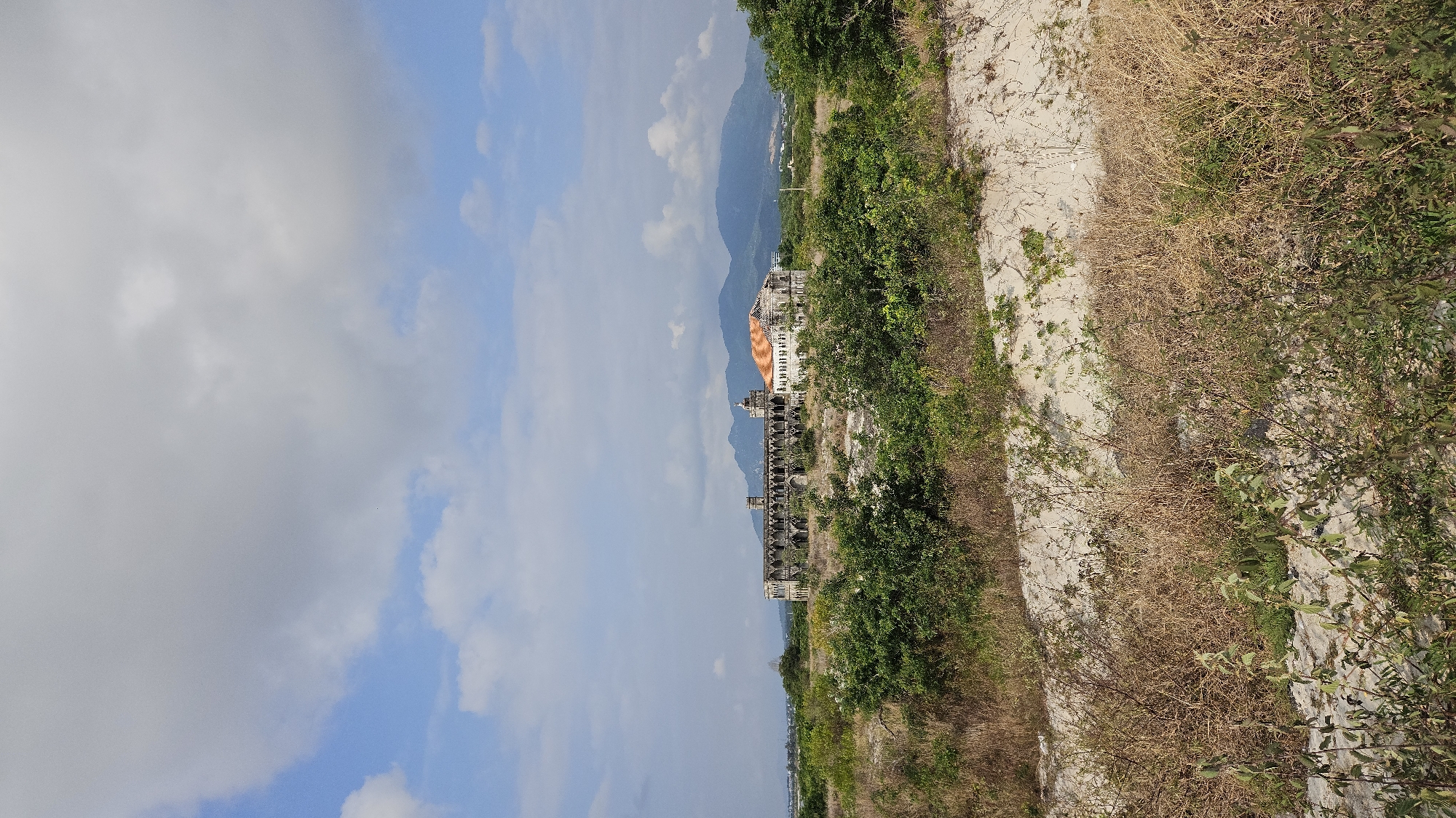 The image shows a ruined, ornate building with an orange roof in Cam Ranh, Vietnam. The structure stands in a desolate area overgrown with dry vegetation and some green foliage. Mountains are visible in the background under a cloudy blue sky. The perspective is slightly tilted.