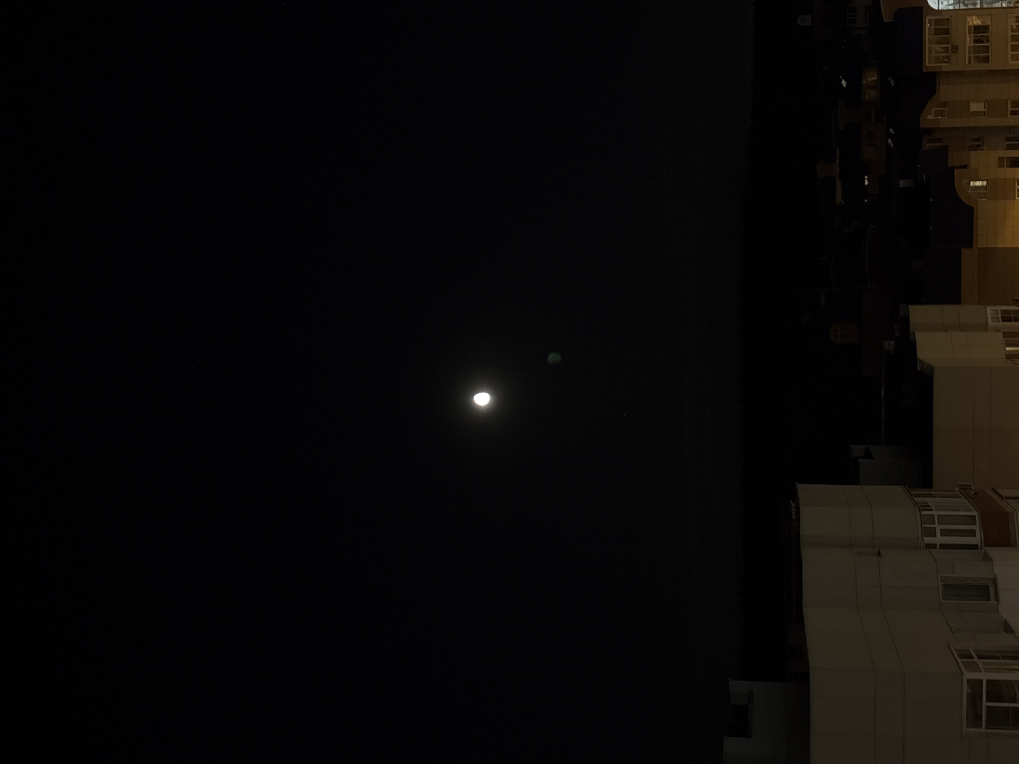 The image depicts a nighttime cityscape, likely from an elevated perspective, looking out over buildings in Balashikha, Russia. A bright moon is visible in the dark sky, with a slight green lens flare or atmospheric effect beneath it. In the foreground, the tops of several residential buildings are illuminated by artificial light, showcasing balconies and windows. The overall scene suggests a quiet, residential area at night.