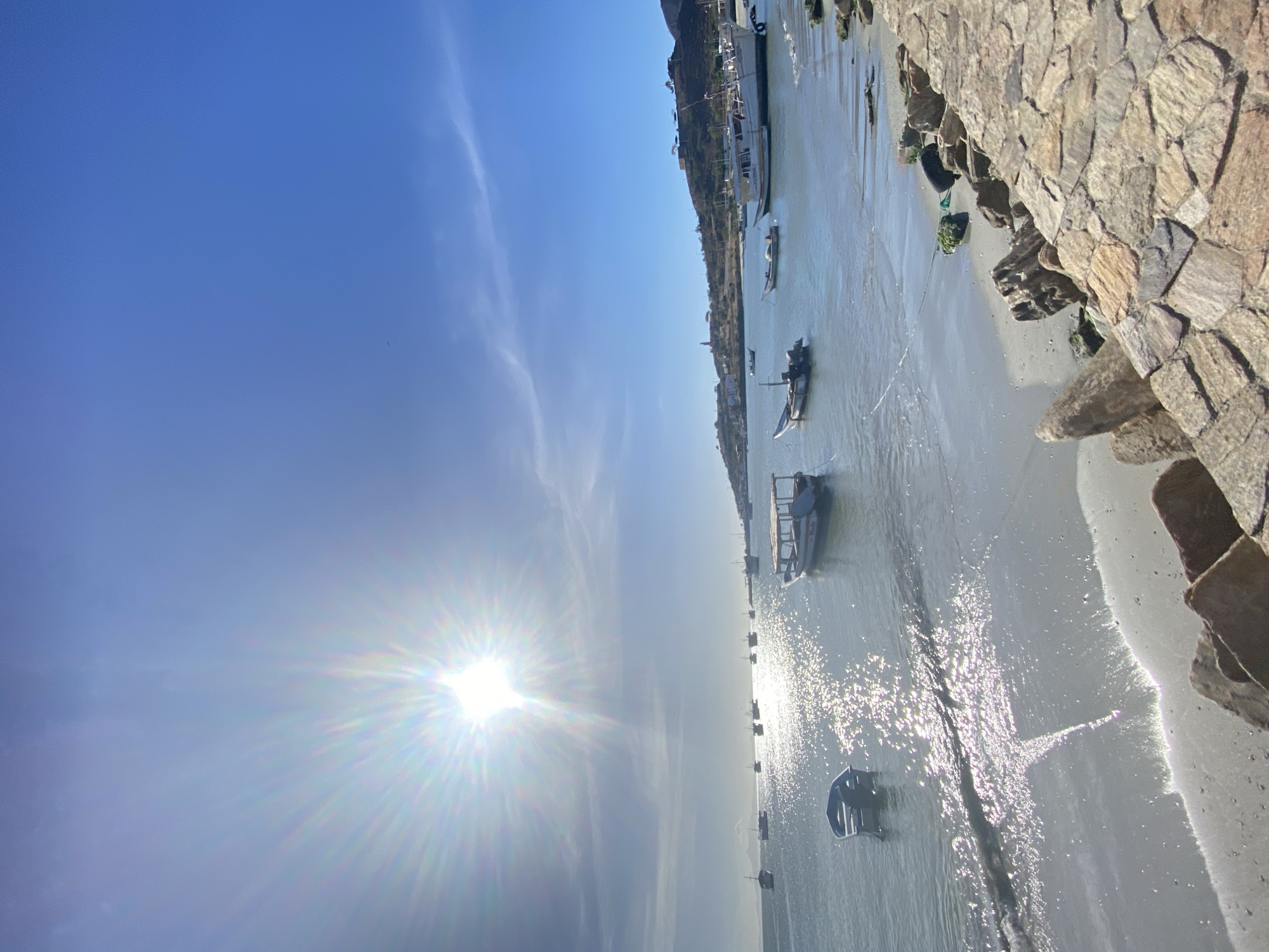 This image depicts a sunlit coastal bay in Juan Griego, Venezuela. The sky is clear blue with sparse, wispy clouds, and a brilliant, starburst sun occupies the upper left quadrant, casting strong reflections across the water. The bay contains approximately 15 to 20 moored or anchored boats of varying sizes, primarily small to medium fishing vessels, some with canopies or visible outboard motors. The water surface exhibits significant sparkling glare from the sun. The immediate foreground on the right features a constructed stone-paved embankment or pathway composed of irregular, flat stones. Below this is a light grey sandy beach with gentle waves reaching the shore. The background consists of dry, undulating hills rising from the water, dotted with structures including a visible white church tower and what appears to be a distant lighthouse. No human activity or individuals are discernible within the frame.