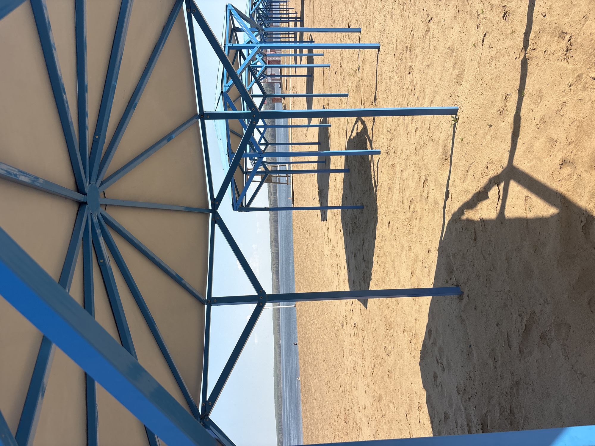The image shows a row of sunshades on a sandy beach in Chaikovsky, Russia. The sunshades have blue metal frames and light-colored fabric tops. The beach is located next to a body of water, possibly a lake or river. The sun is shining, casting shadows of the sunshade frames onto the sand. There are no people visible in the image. The scene appears calm and empty.