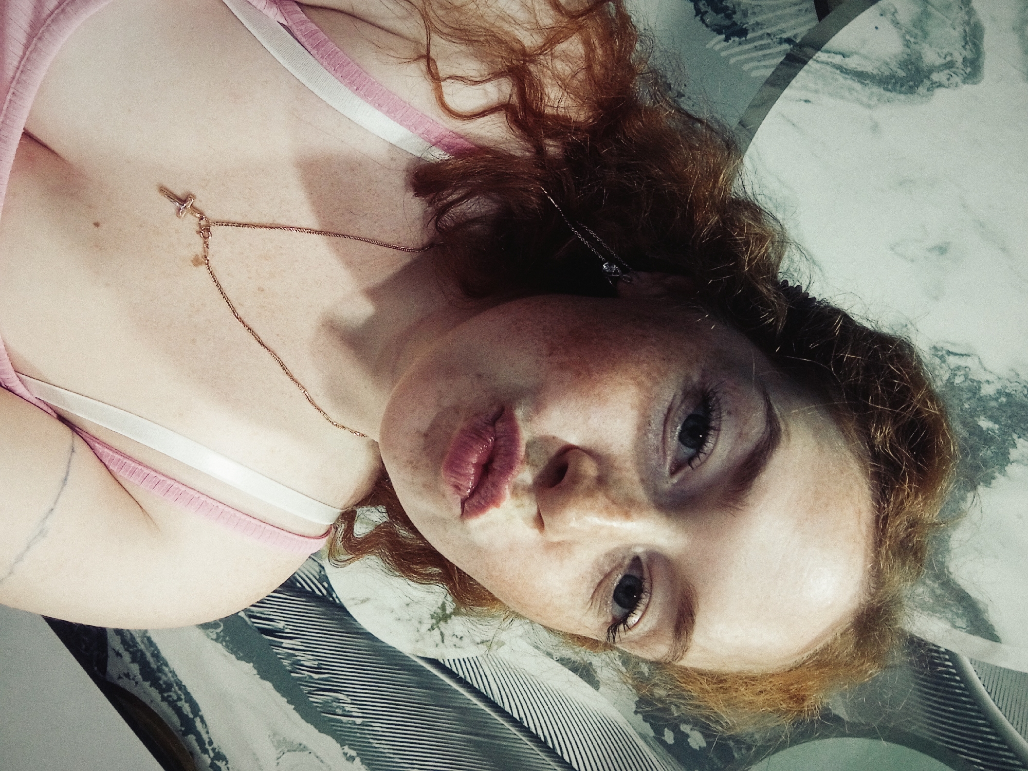 A close-up photograph shows a light-skinned individual with red hair and freckles, lying on what appears to be a patterned surface. They are wearing a pink tank top with a white strap. A gold necklace with a cross pendant rests on their chest. The individual is looking into the camera with a slight pout. The background appears to be a surface with a marble-like or textured pattern. The location is not evident from the image itself; the claim that the image was taken in Yurgamysh, Russia, cannot be verified.