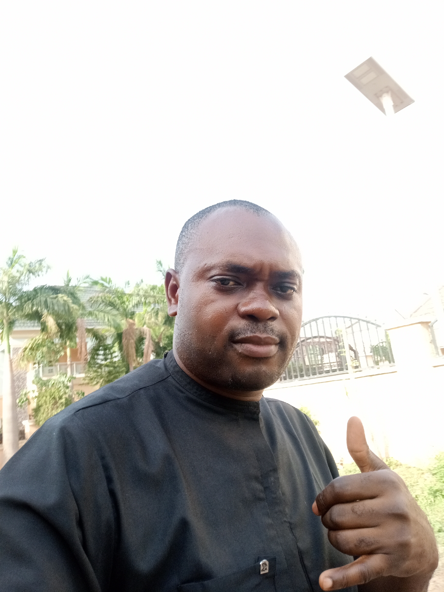 The image is a bright, outdoor selfie of a man. He has dark skin, short dark hair, dark eyes, and a trimmed mustache and beard. He is wearing a plain black, short-sleeved collared shirt. On the left chest pocket of his shirt, there is a small, silver-colored emblem that depicts a stylized figure, though specific details or text are not clearly discernible. The man is looking directly at the camera with a neutral to slightly serious expression and is giving a "thumbs up" gesture with his right hand, positioned in the lower right foreground.
The background is brightly illuminated by natural light, suggesting it is daytime with a clear, possibly sunny, sky. On the left side of the frame, several tall palm trees with green fronds are visible, along with glimpses of a building behind them, suggesting a residential structure. To the right, a dark, ornate metal gate or fence forms part of the mid-ground, with more distant buildings behind it. In the upper right corner, a white rectangular object, resembling a solar panel or an outdoor light fixture, is visible against the bright sky. The setting appears to be a residential or semi-urban environment in Gwarinpa, Nigeria. No legible text is visible within the image.