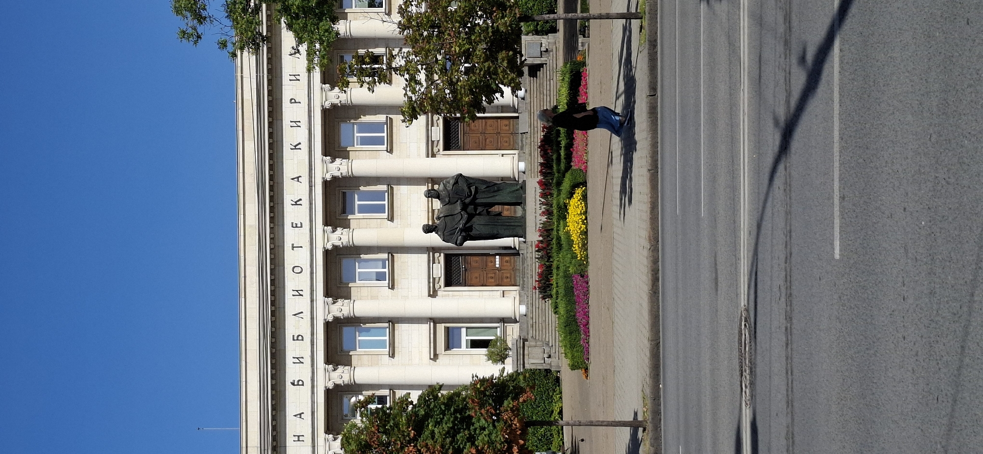 The image shows the Ivan Vazov National Library in Sofia, Bulgaria. The facade of the building features a series of neoclassical columns and an inscription in Cyrillic letters that reads "НАЦИОНАЛНА БИБЛИОТЕКА КИРИЛ И МЕТОДИЙ" (National Library "St. Cyril and Methodius"). In front of the entrance are two bronze statues. A neatly manicured flower bed with vibrant pink, yellow, and red flowers is visible below the statues. A paved walkway leads from the library entrance towards a street in the foreground. A woman wearing a dark top and a blue skirt is walking on the paved walkway, casting a shadow on the ground. The street is asphalt, marked with white lines, and a manhole cover is visible. Trees with green and reddish foliage are situated to the left and right of the building. The sky is clear blue, indicating a sunny day.