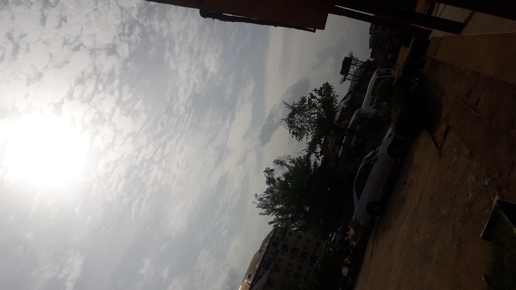 The image shows a street scene in Onitsha, Nigeria. Several cars are parked along a concrete path beside the curb. A multi-story building is visible in the background with a water tank or similar industrial equipment visible. The sky is bright with scattered cloud cover. Trees are planted along the curb. Parts of a building are visible overhead. No people are clearly visible.