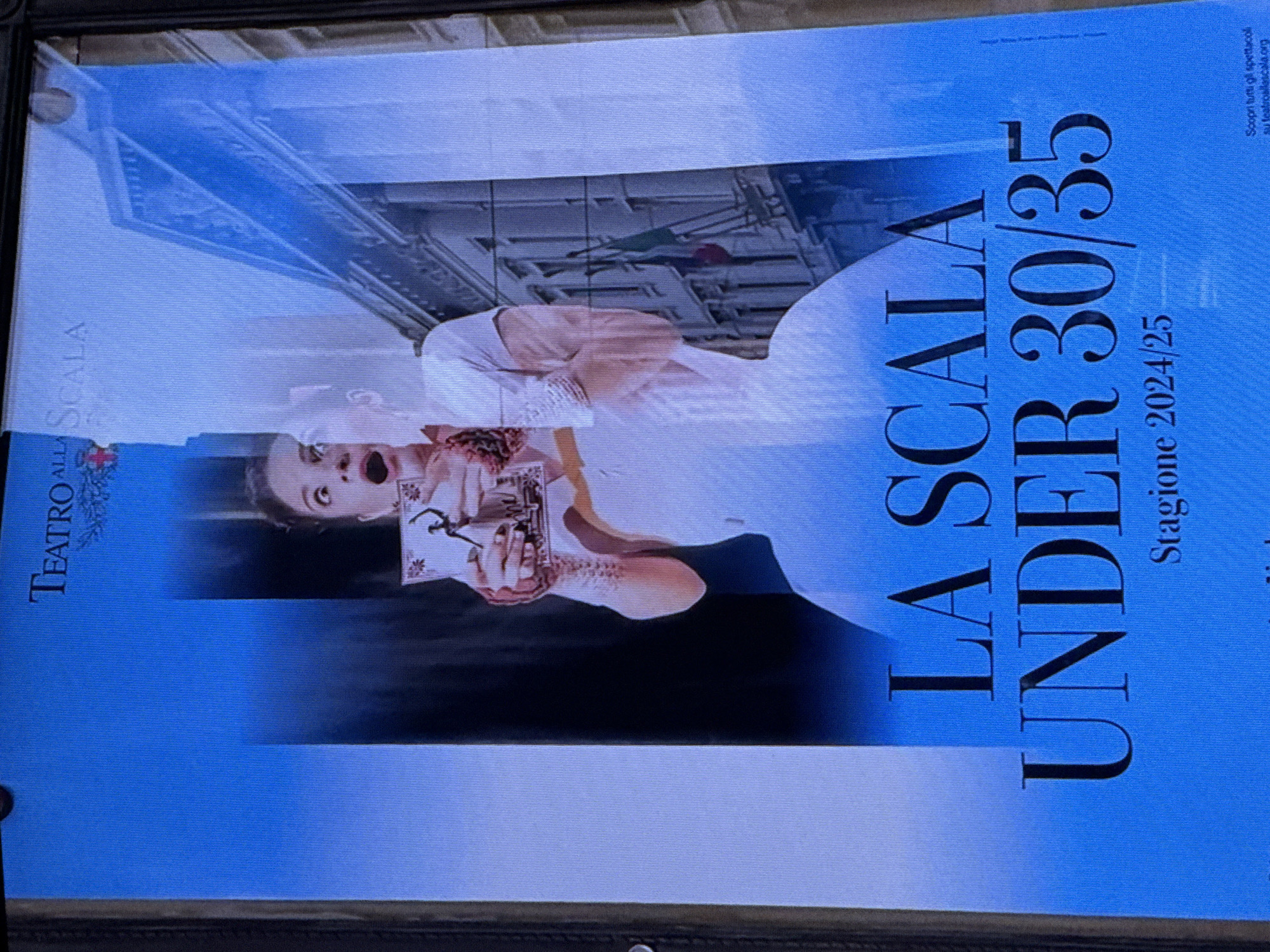 The image is a photograph of a poster for "Teatro alla Scala" for the "UNDER 30/35" season in 2024/25. The poster features a woman in a white dress with her mouth open in surprise as she holds up a framed picture of a dancer. Behind her is a stylized depiction of a building, possibly the Teatro alla Scala itself. The poster is predominantly blue with large black text indicating the season and age group. There is no indication of the city of Dubai or the United Arab Emirates in this image.