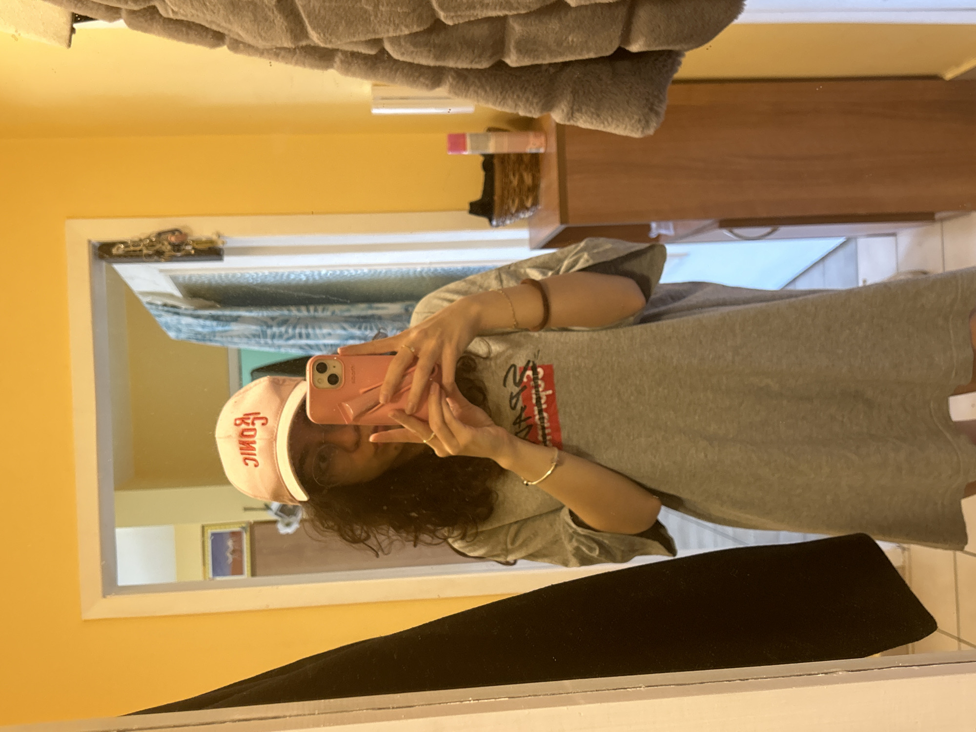 A person is taking a mirror selfie inside a room. The room appears to be a bathroom with yellow walls, a white-framed mirror, and a wooden shelf. The person is wearing a gray t-shirt, a pink "Iconic" baseball cap, and jewelry on their wrists. The reflection in the mirror shows a framed picture on the wall and a view of a building exterior. The location in the city of Sofia, Bulgaria, cannot be determined from the image.