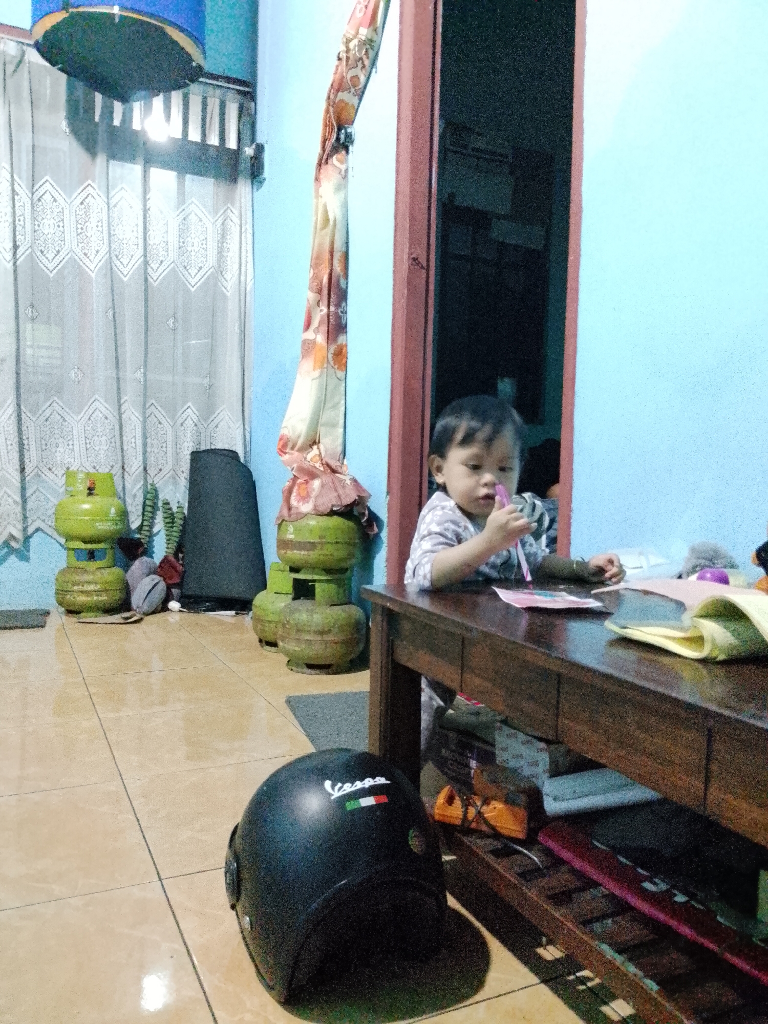 A young child is seated at a wooden table, holding a pink object to their mouth. The child is wearing a light-colored shirt with a pattern. The table is dark wood and has several items on it, including papers and what appears to be a yellow notebook or folder. To the left of the table, there are two stacks of green propane gas cylinders. Behind the cylinders is a dark conical object and some green decorative plants. A patterned curtain hangs from a rod near a doorway. The room has light blue walls and tiled flooring. On the floor in the foreground, to the left, is a black helmet with "Vespa" written on it in white, accompanied by the Italian flag colors. The overall scene appears to be an indoor domestic setting, possibly in Indonesia, given the location context. The lighting suggests it could be daytime, with light coming from a window covered by sheer curtains on the left side of the image. The child's expression is neutral.
