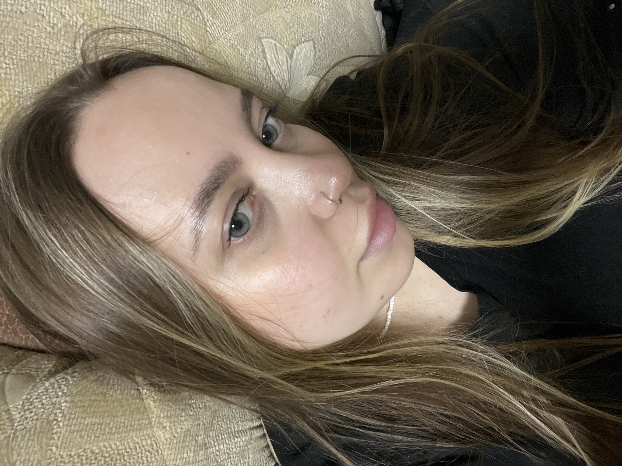 A close-up, eye-level shot shows a fair-skinned woman with light brown to blonde hair lying down, possibly on a bed or couch, as suggested by the patterned fabric underneath. Her head is tilted slightly, and she is gazing towards the camera with light-colored eyes. She has a small nose ring. Her clothing appears to be a dark color. The background is blurred, but it seems to consist of more of the dark-colored material. The location in the city of Novopokrovskaya, Russia is unknown.