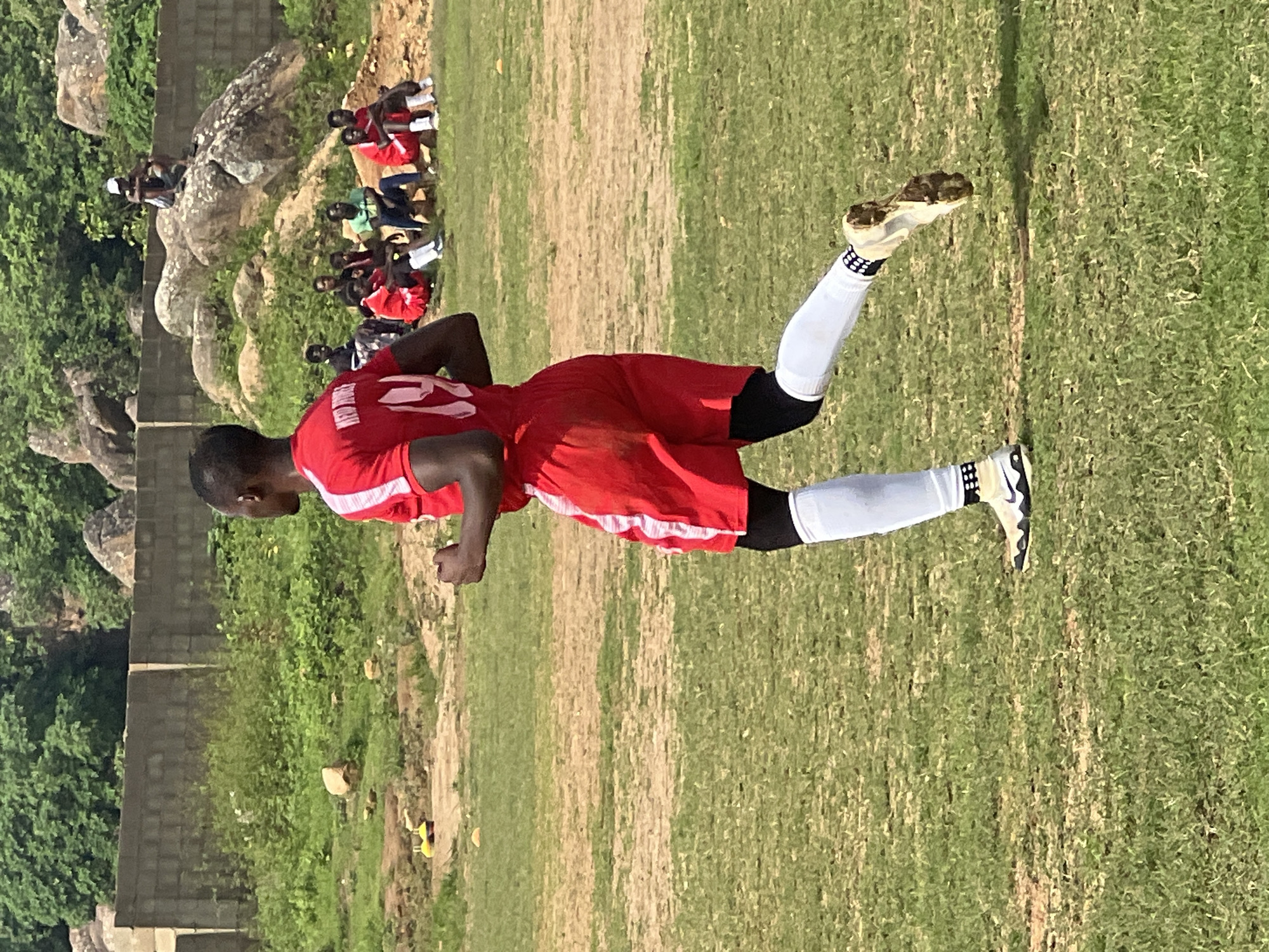 In the city of Jos, Nigeria, a male soccer player is captured in a mid-stride motion on a grassy field, his body angled away from the camera and to the right. He wears a red jersey with white trim and the number "14" in white on the back, red shorts, black compression sleeves on his lower legs, and white socks with a black patterned band at the top. His cleats are white with black accents and are visibly soiled with dirt. In the background, a rough stone wall separates the field from a natural landscape featuring large boulders and dense green foliage. A group of spectators, appearing to be young males, are seated on the rocks and the ground beyond the wall, observing the activity on the field. Some are also seated on a raised platform behind them, also watching. A single individual sits atop a prominent boulder in the upper background. The lighting suggests it is a bright, sunny day.