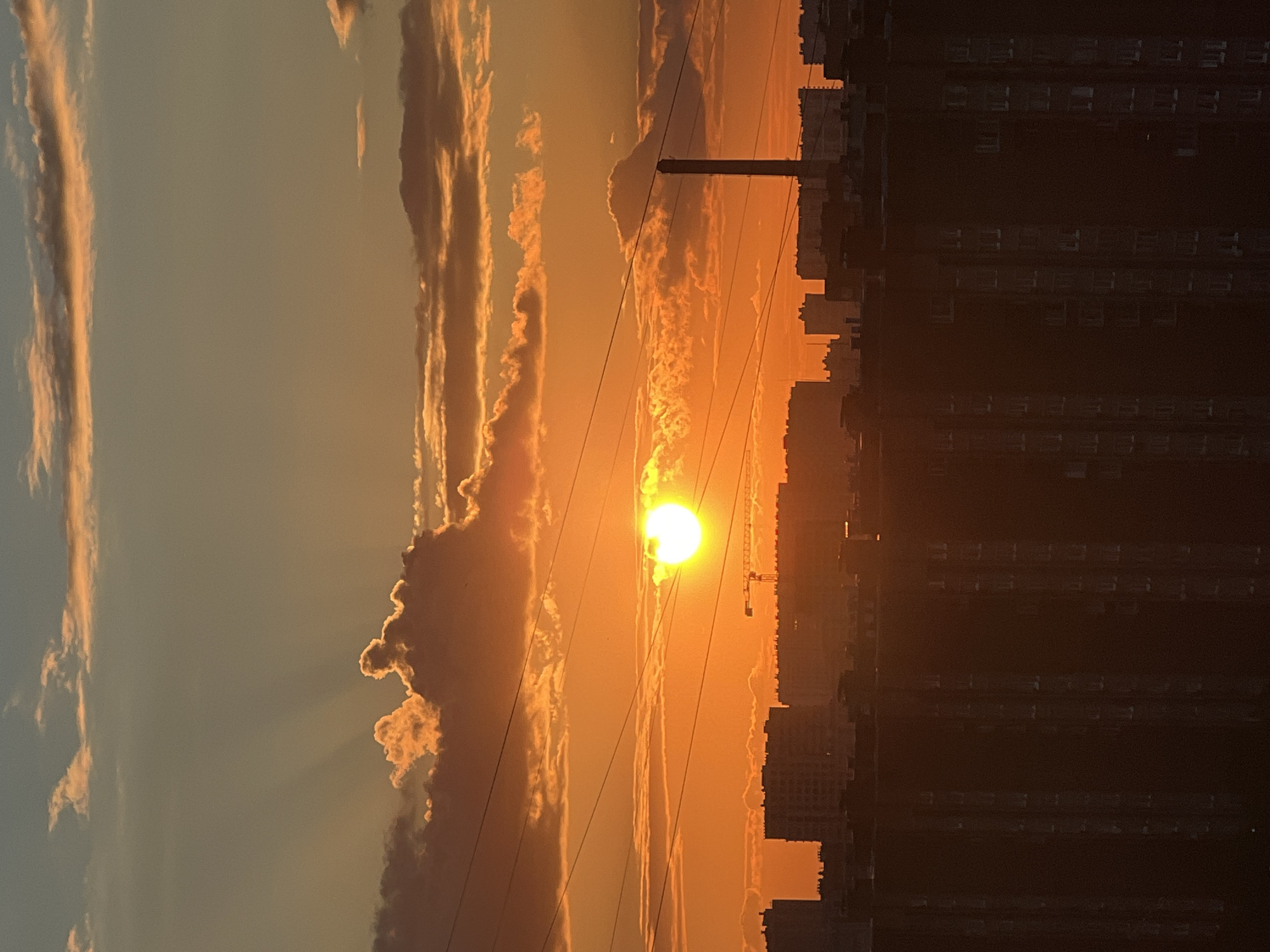 This media file captures a sunset over an urban landscape, likely in Russia. The sun, a bright orb of orange, is positioned in the center of the frame, casting a warm glow across the sky and illuminating scattered clouds. The clouds are tinged with shades of orange, pink, and yellow, with some appearing dark and dramatic against the vibrant sky.
In the foreground, silhouetted against the setting sun, are power lines and a utility pole, indicating an urban or semi-urban environment. To the right of the sun, the dark, angular shapes of apartment buildings rise up, their windows visible as faint rectangles. The buildings are densely packed, suggesting a city setting. The overall atmosphere is serene and picturesque, evoking a sense of peace and the end of the day. The time of day is clearly sunset, and the weather appears clear with some cloud formations. No people are visible in the image, and there is no visible text.