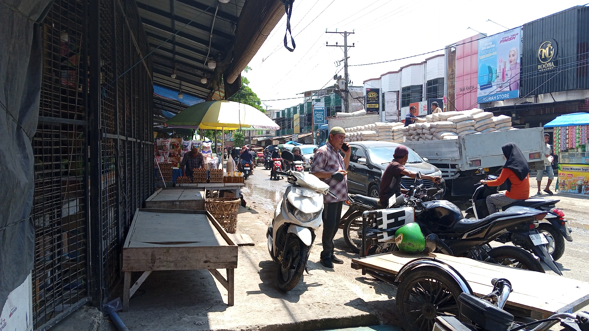 This image depicts a street scene in Lhokseumawe, Indonesia, likely a market area. The foreground features a covered market stall with a caged structure and wooden tables. Several motorcycles are parked, including a white scooter in the center and a black motorcycle on the right with a wooden trailer attached.
In the mid-ground, a man in a checkered shirt and a skullcap is talking on a mobile phone, holding what appears to be a small item in his other hand. A truck loaded with sacks of what looks like cement or similar material is parked on the road, with a woman wearing a black headscarf and an orange shirt riding a motorcycle next to it. Another person, a man in a dark shirt and maroon cap, is seated on a motorcycle.
The background shows a busy street with more motorcycles, cars, and buildings. A prominent billboard with an advertisement for Wardah is visible on one of the buildings. The environment appears to be a sunny day, with a mix of commercial establishments and open market stalls. The road surface is wet in places, suggesting recent rain or a cleaning activity. Several people are present, engaged in various activities like selling, commuting, or interacting.