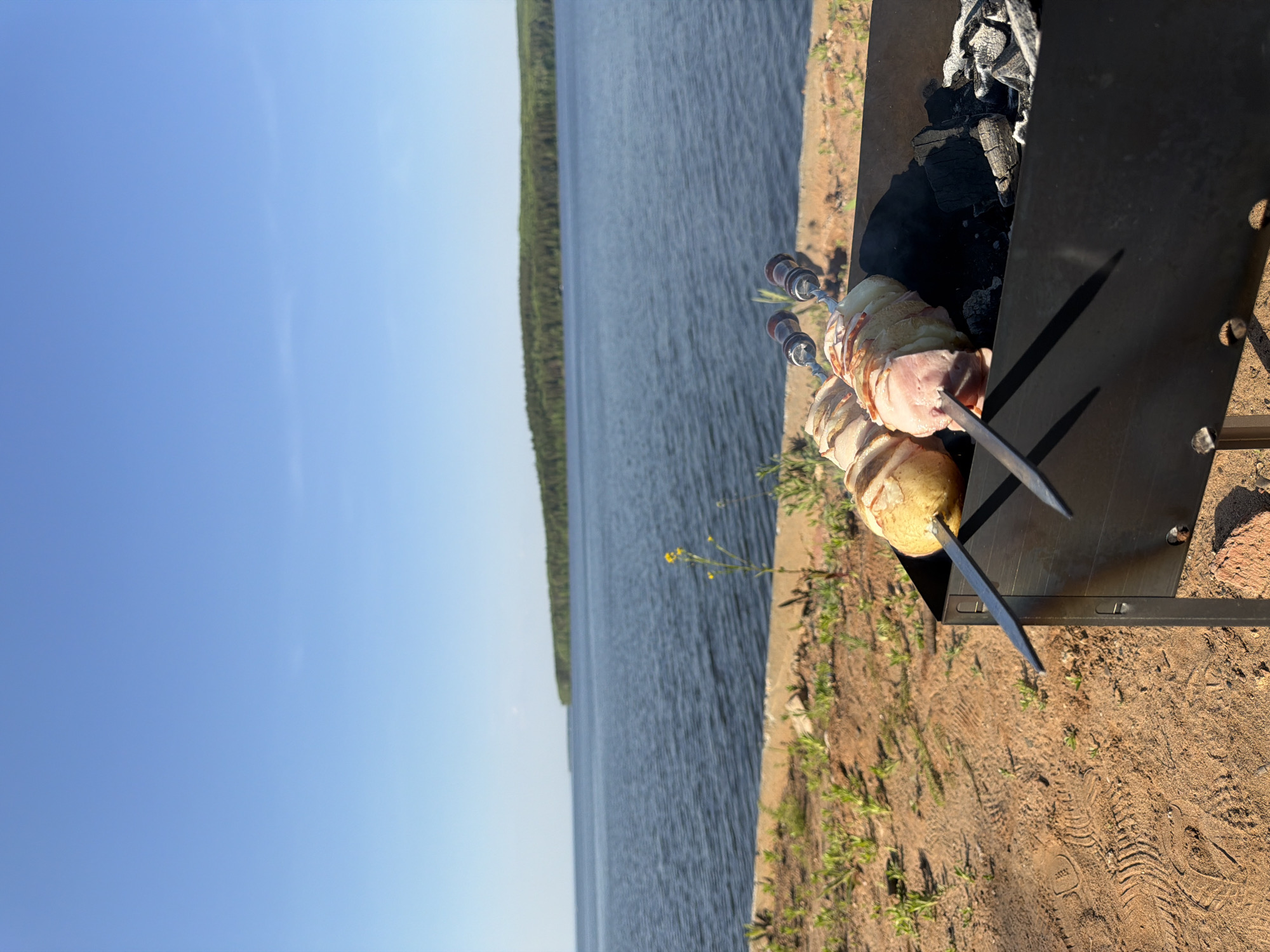 The image shows a barbecue setup outdoors, likely near a body of water, possibly the Kama Reservoir in Chaikovsky, Russia. The focus is on skewers with food, likely potatoes wrapped with bacon, positioned over a charcoal grill. The grill sits on a sandy or earthen surface with sparse vegetation. In the background, a body of water extends towards a treeline under a clear blue sky. The photo appears to be taken during daylight, with natural light illuminating the scene. The overall impression is one of a leisure activity in a natural environment.
