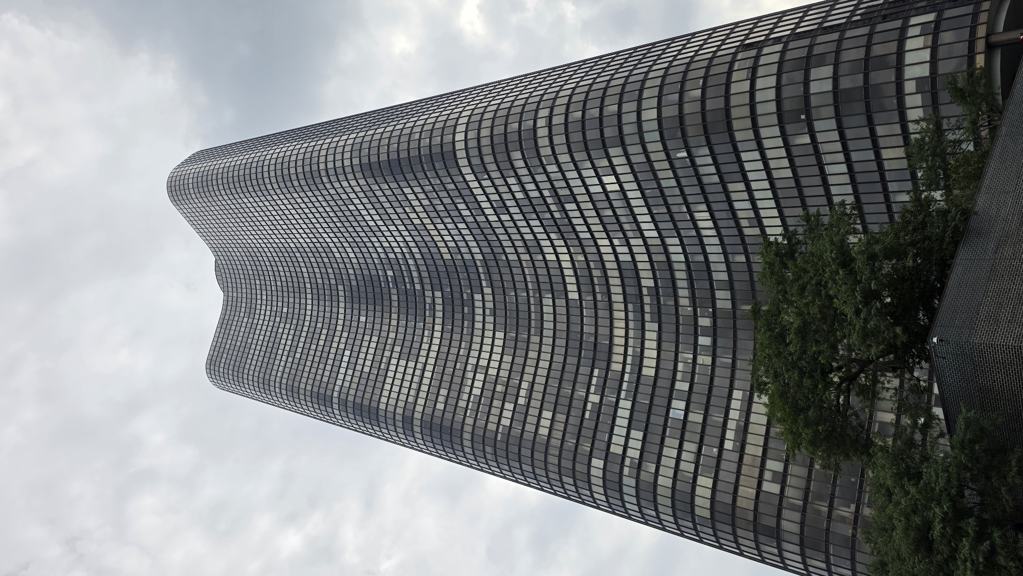 The image shows a low-angle, wide shot of the Marina City towers in Chicago, Illinois, United States of America. The iconic twin residential skyscrapers, known for their distinctive sail-like, curved exteriors, dominate the frame under a cloudy sky. The buildings are clad in dark glass and steel, with a grid-like pattern of windows reflecting the overcast conditions. In the foreground, a dense canopy of green trees is visible, partially obscuring the lower levels of the buildings. A dark brick structure with a visible security camera is also present at the base of the towers. The perspective emphasizes the immense height and unique architectural design of the Marina City towers.
