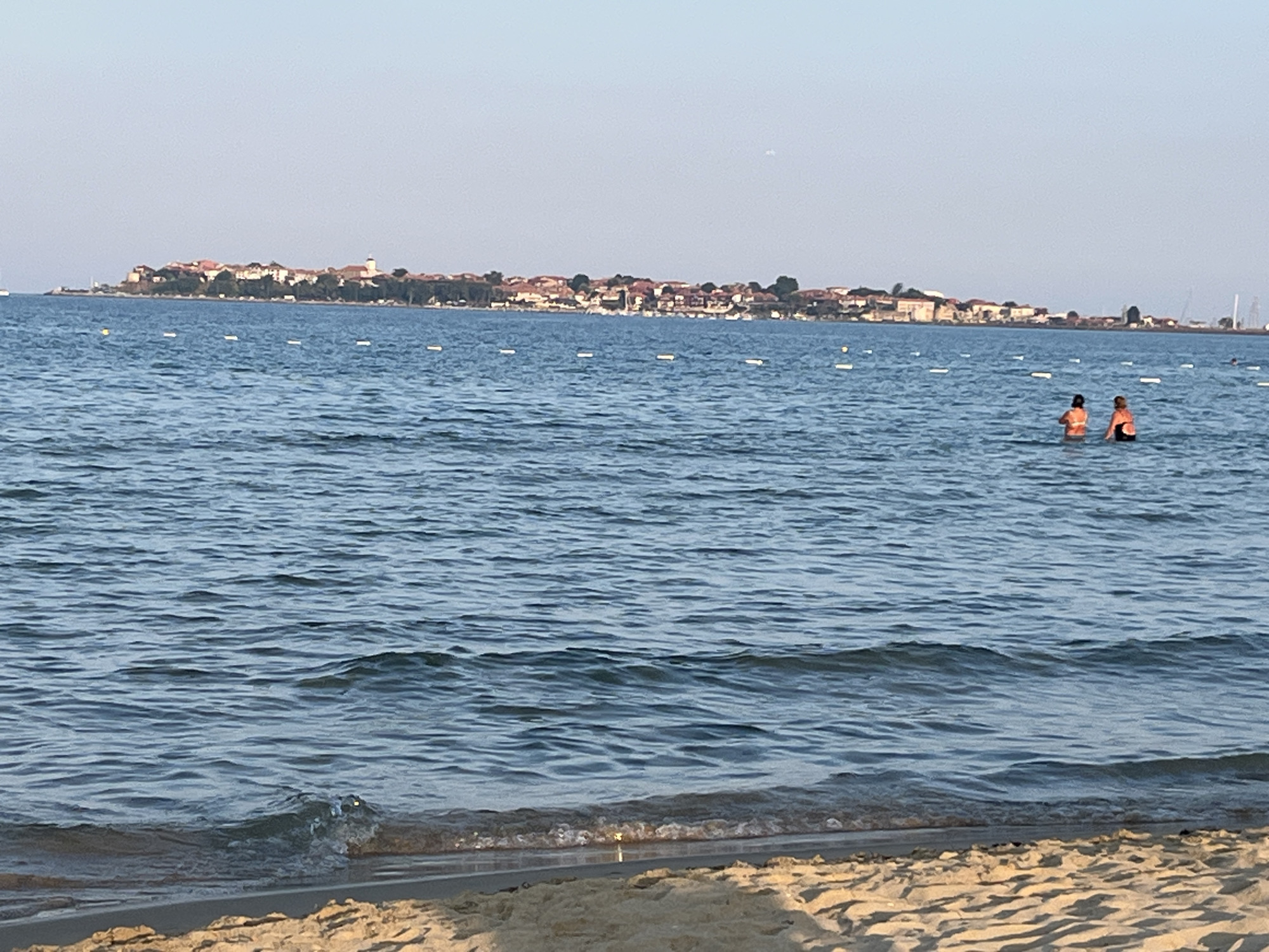 The image depicts a tranquil scene at the beach in Nesebar, Bulgaria. In the foreground, the sandy shore meets the gentle waves of the Black Sea. Two individuals, wearing life vests, are visible wading in the water. In the background, the picturesque old town of Nesebar, a UNESCO World Heritage site, is situated on a peninsula, characterized by its red-tiled roofs and historic buildings. The sky above is a clear, pale blue, and a few buoys are dotted across the water, marking the bathing area. The overall atmosphere is serene, capturing a typical summer day at the Bulgarian coast.