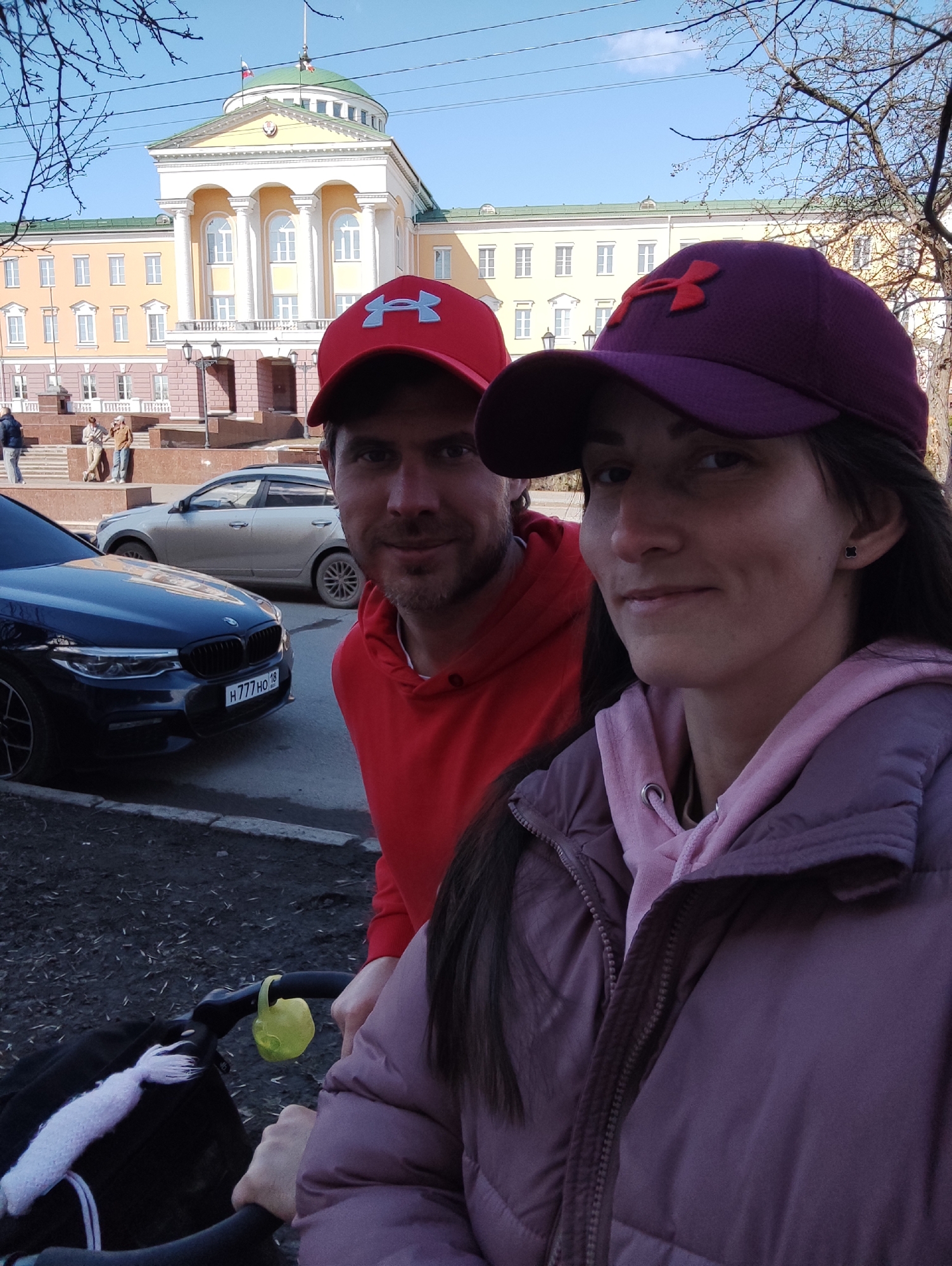 A couple poses in front of the State Council of the Udmurt Republic in Izhevsk, Russia. The woman in the foreground wears a purple Under Armour cap and puffer jacket over a pink hoodie. A stroller is partially visible in front of her. To her left and slightly behind, a man wears a red Under Armour cap and hoodie. A black BMW car is parked on the street to their left, with the license plate number H777HO 18 visible. In the background, the State Council building has a pale yellow facade, white columns, and a green dome with a flag. Other cars and pedestrians are visible in the background. Bare tree branches frame the top of the image against a clear blue sky.