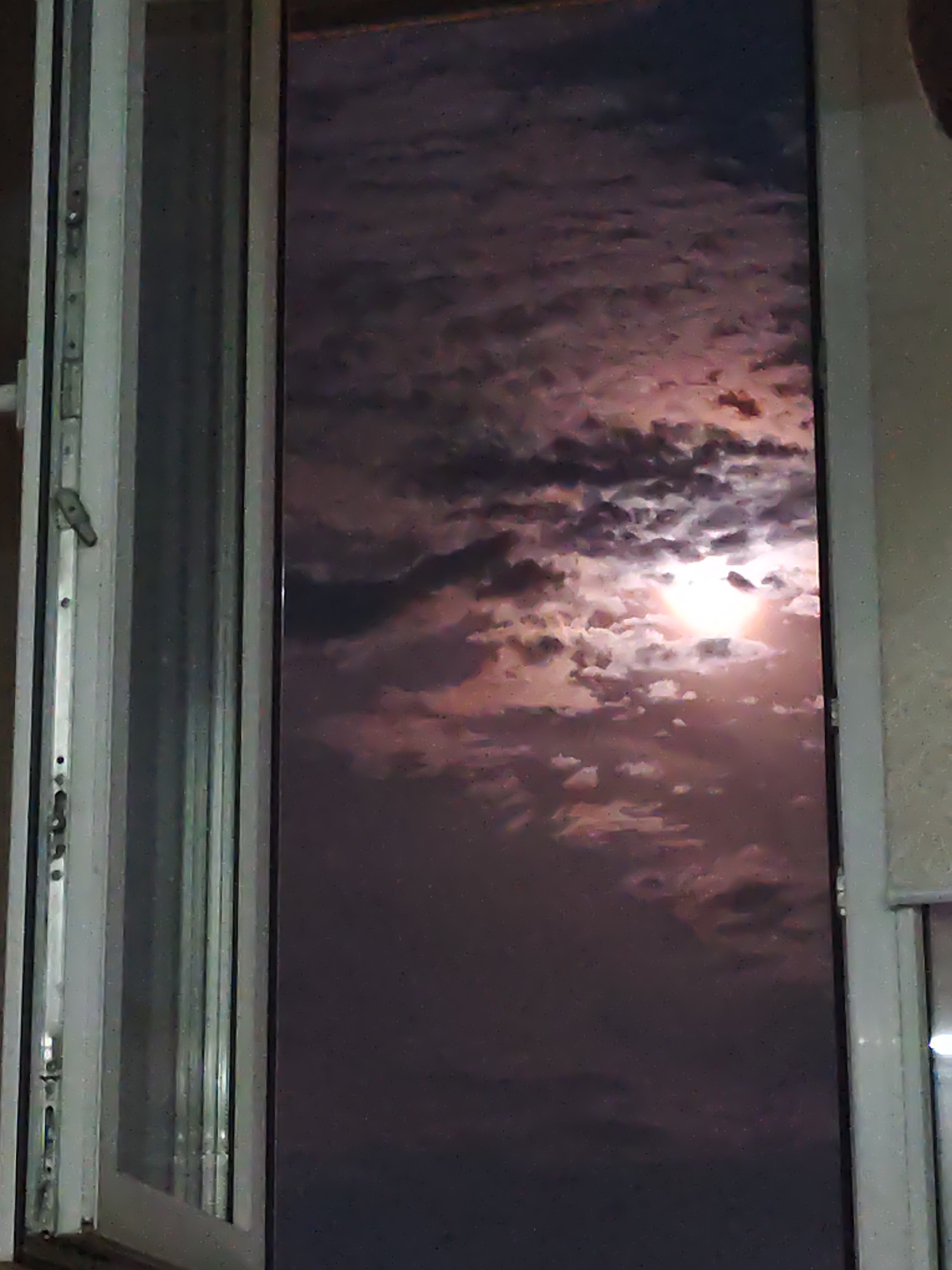 The image shows a night sky with the moon and clouds, viewed through a window. The moon is bright and illuminated, surrounded by pink and purple clouds. The window appears to be part of a residential building, with a frame and glass visible. The overall scene suggests a peaceful and somewhat romantic atmosphere. There are no people or specific landmarks that would allow for a precise location within Petropavl, Kazakhstan, to be determined from the image alone.