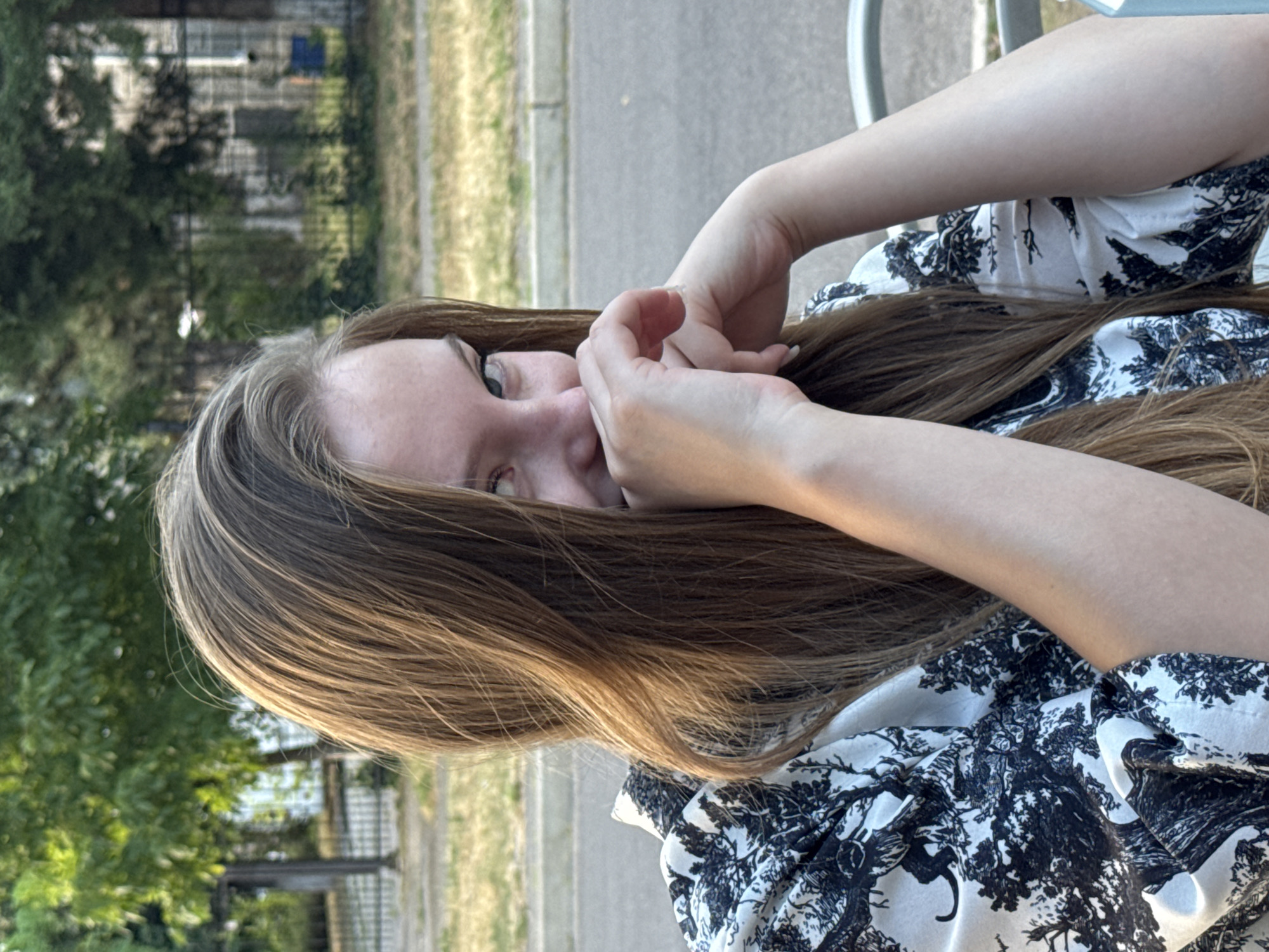 A young woman with long, brown hair is pictured from the chest up, seated outdoors. Her face is partially obscured by her hands, which are clasped near her mouth, and her hair falls across her left side. She is wearing a white blouse with a black, intricate pattern of trees and possibly animals. The background suggests an urban or suburban setting with trees, a fence, and a road visible. The lighting indicates it is daytime, likely late afternoon given the warm tones. The scene appears to be in Tiraspol, Moldova, with the presence of characteristic urban foliage and infrastructure.