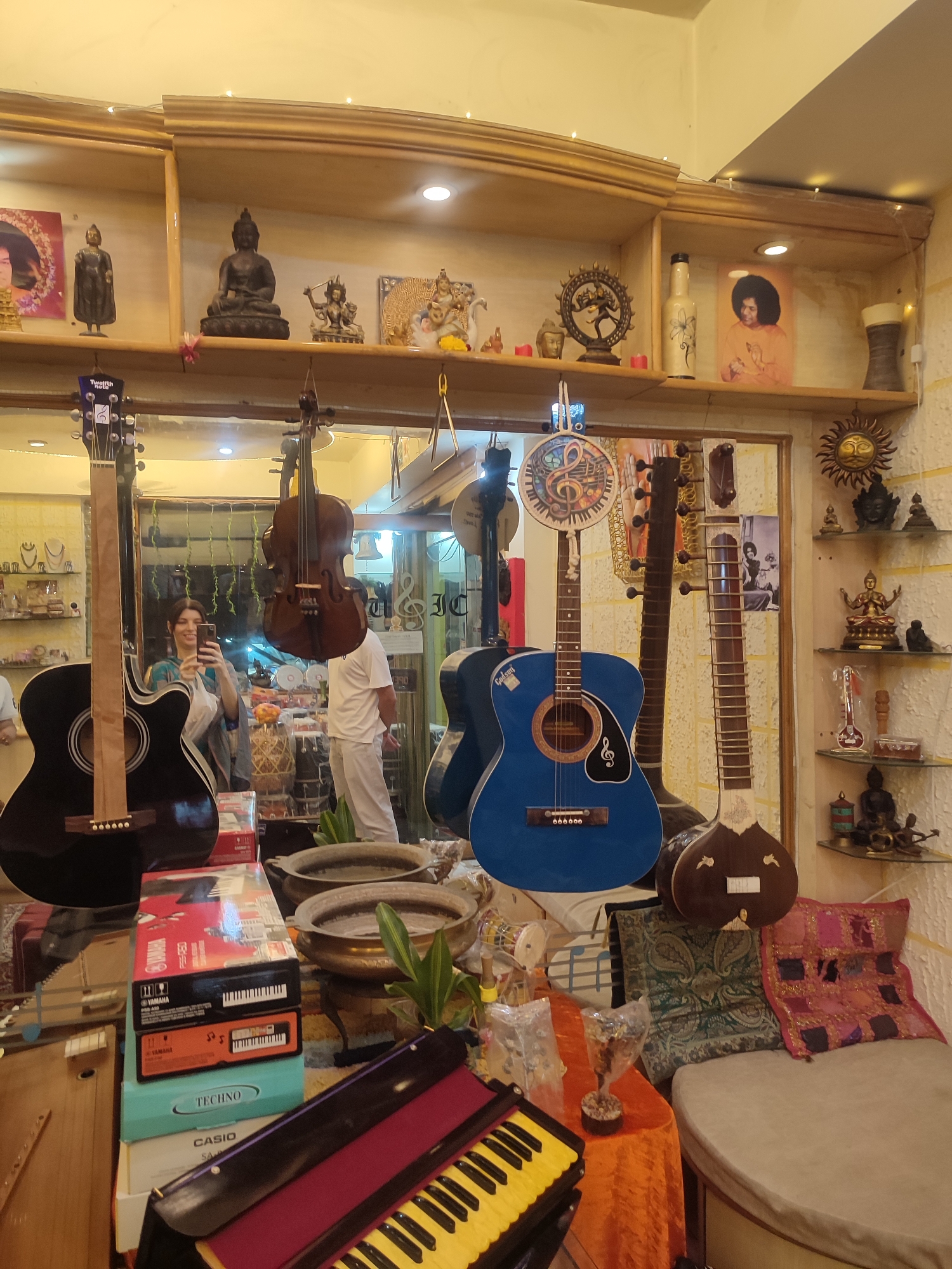 This image captures the interior of a shop in Puttaparthi, India, specializing in musical instruments and spiritual artifacts. The scene is dominated by a large mirror reflecting much of the store and a woman taking a photo with her phone. She is smiling and wearing a blue and grey patterned garment. Another person, partially visible in the reflection, wears white clothing.
Numerous musical instruments are on display. In the foreground, a black acoustic guitar stands upright on the left. A bright blue acoustic guitar, possibly branded "GIBSON" (partially visible), hangs near the center, decorated with a circular motif featuring a musical note. To its right, a dark brown sitar is prominently displayed. Hanging above are a dark wooden string instrument resembling a cello or violin, and another string instrument. In the foreground on a table, a yellow and black keyboard (harmonium) is visible, along with several boxes for electronic keyboards, including "Yamaha PSR-A50," "TECHNO," and "CASIO SA-46."
The shop's shelves are laden with an array of spiritual and decorative items. These include various statues of Buddha in different poses, figures of Hindu deities such as Shiva Nataraja and what appears to be Ganesha, and framed images of spiritual leaders like Sai Baba. Other decorative elements include bottles, small pots, and a stylized sun motif on the wall. Small fairy lights adorn the top of the shelving units. Colorful, embroidered cushions are arranged on a low seating area on the right.
The overall setting suggests a vibrant retail space, likely catering to tourists or devotees in Puttaparthi, offering both musical instruments and items of spiritual significance. The woman's smile indicates a pleasant shopping experience. The visible text on the guitar headstock reads "Twelfth note," and a reversed "DESIGN" sign is visible in the mirror. The artificial lighting suggests it could be any time of day, but the indoor setting is the primary focus.