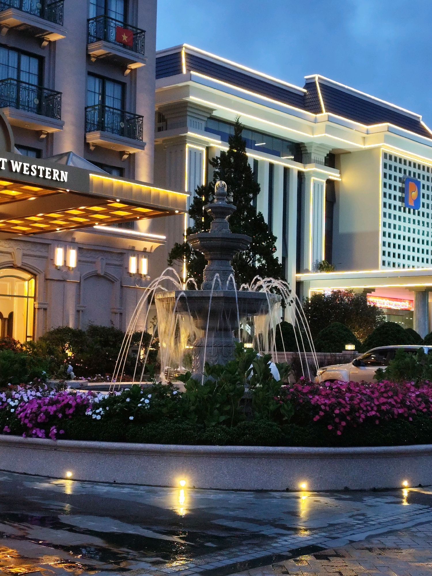 The image shows a fountain in front of a hotel in Lam Vien - Da Lat, Vietnam. The hotel is lit up at night with bright lights, and the fountain is spraying water. There are also flowers and trees around the fountain. A car is parked in the background. The sky is dark, indicating it is nighttime.