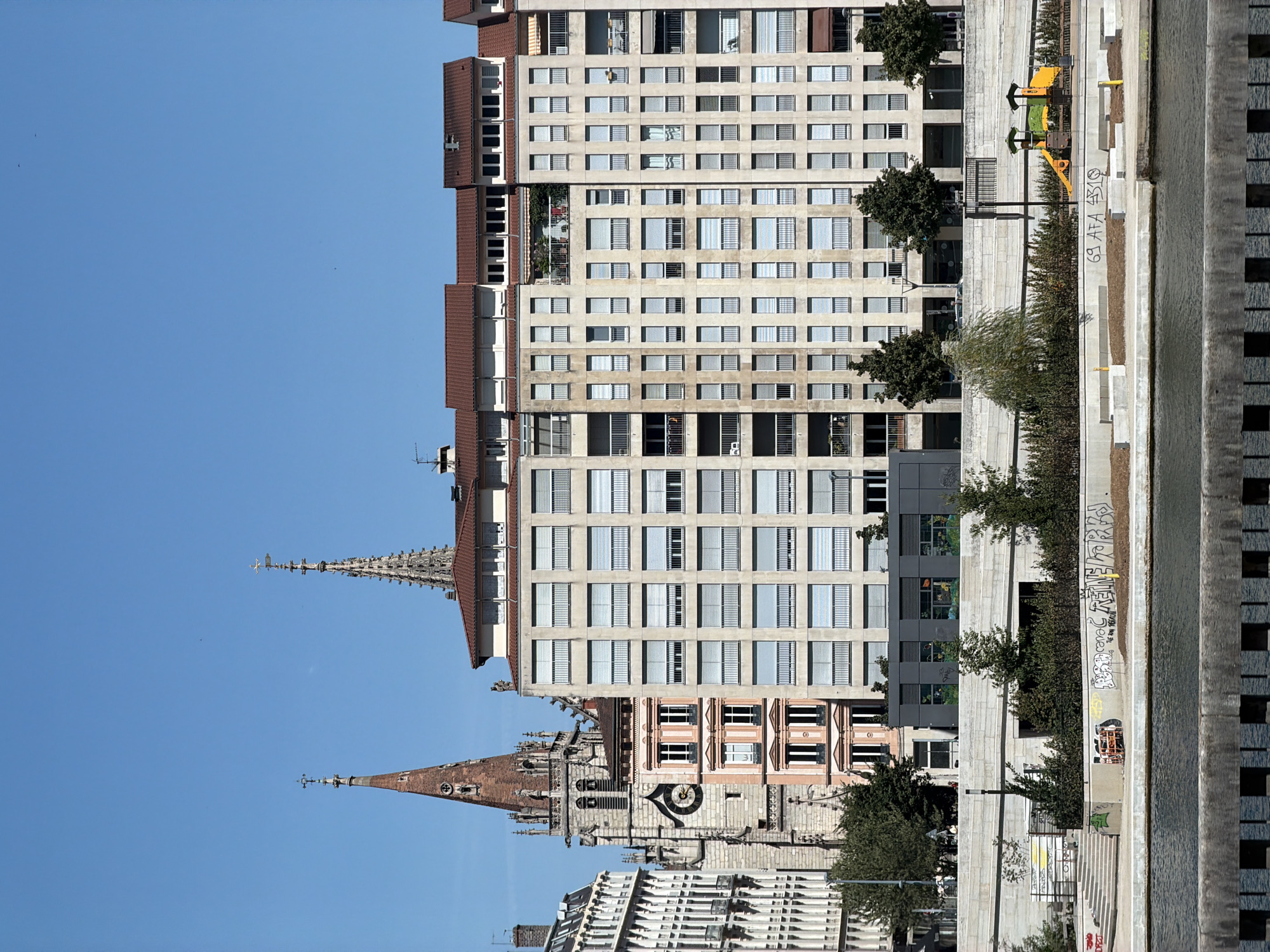 The image depicts a wide shot of a riverfront in Lyon, France. On the left, an ornate church steeple rises above a pale pink building with numerous windows and a prominent clock face. To the right of the church is a modern, multi-story building with a beige facade, rows of windows, and balconies. Further to the right, more of this modern building and another building with a red-tiled roof are visible.

The river occupies the foreground, with a grey, textured barrier along its edge. Behind the river barrier is a concrete promenade or walkway. On this level, there are stairs leading up to the buildings, trees, and graffiti on the concrete walls. To the right of the promenade, a children's playground with yellow and green equipment is visible. The sky above is a clear, bright blue with a few distant birds. The overall scene suggests a sunny day in an urban environment along a waterway.