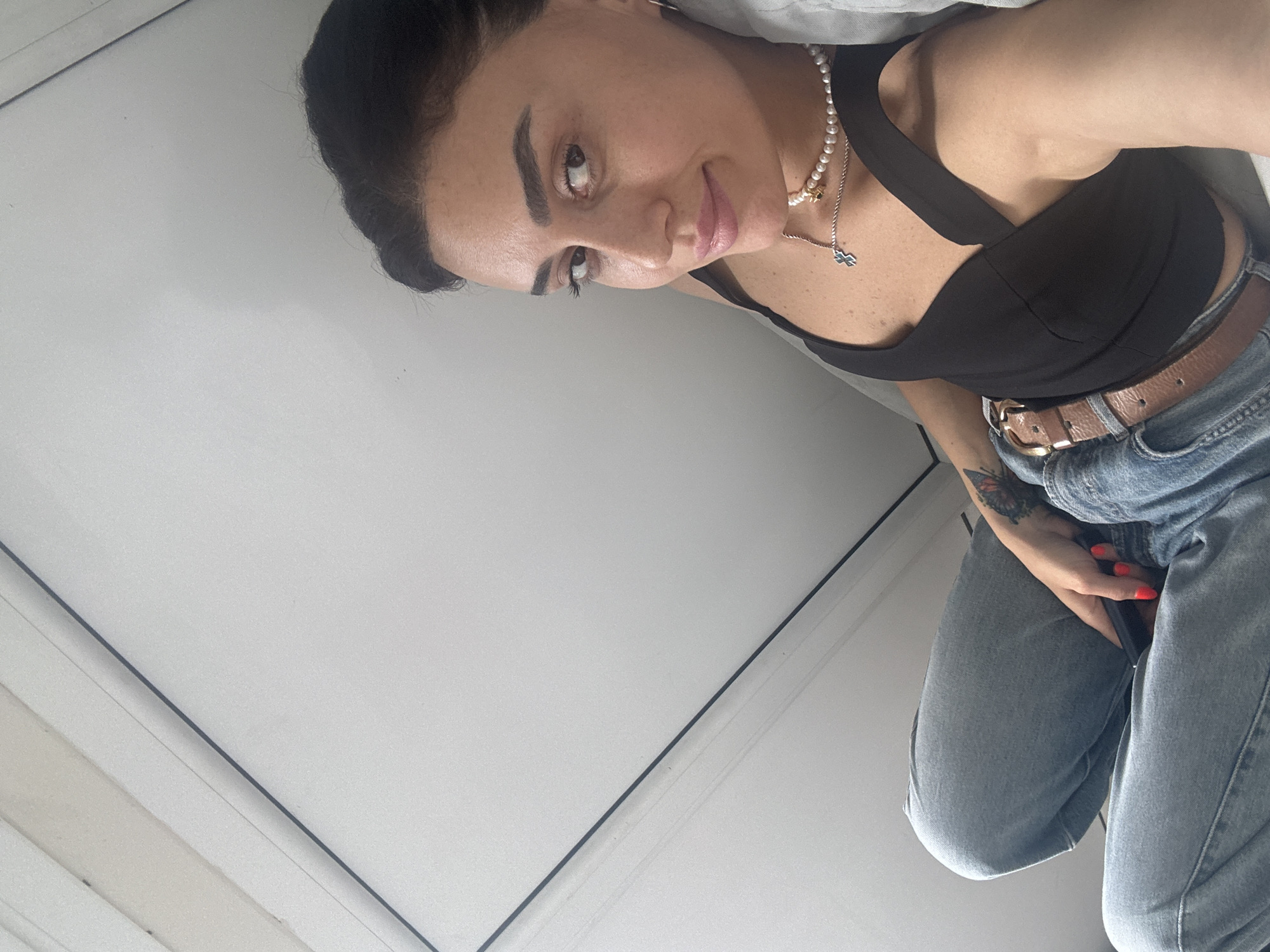 A woman with dark hair and brown eyes is captured in a selfie, lying down with her legs crossed and holding a phone. She is wearing a black top, blue jeans, and a brown belt. A pearl necklace with a silver cross pendant adorns her neck. A butterfly tattoo is visible on her left arm. The background consists of a white wall and ceiling, possibly a room or apartment in Tbilisi, Georgia. The lighting suggests it is daytime.