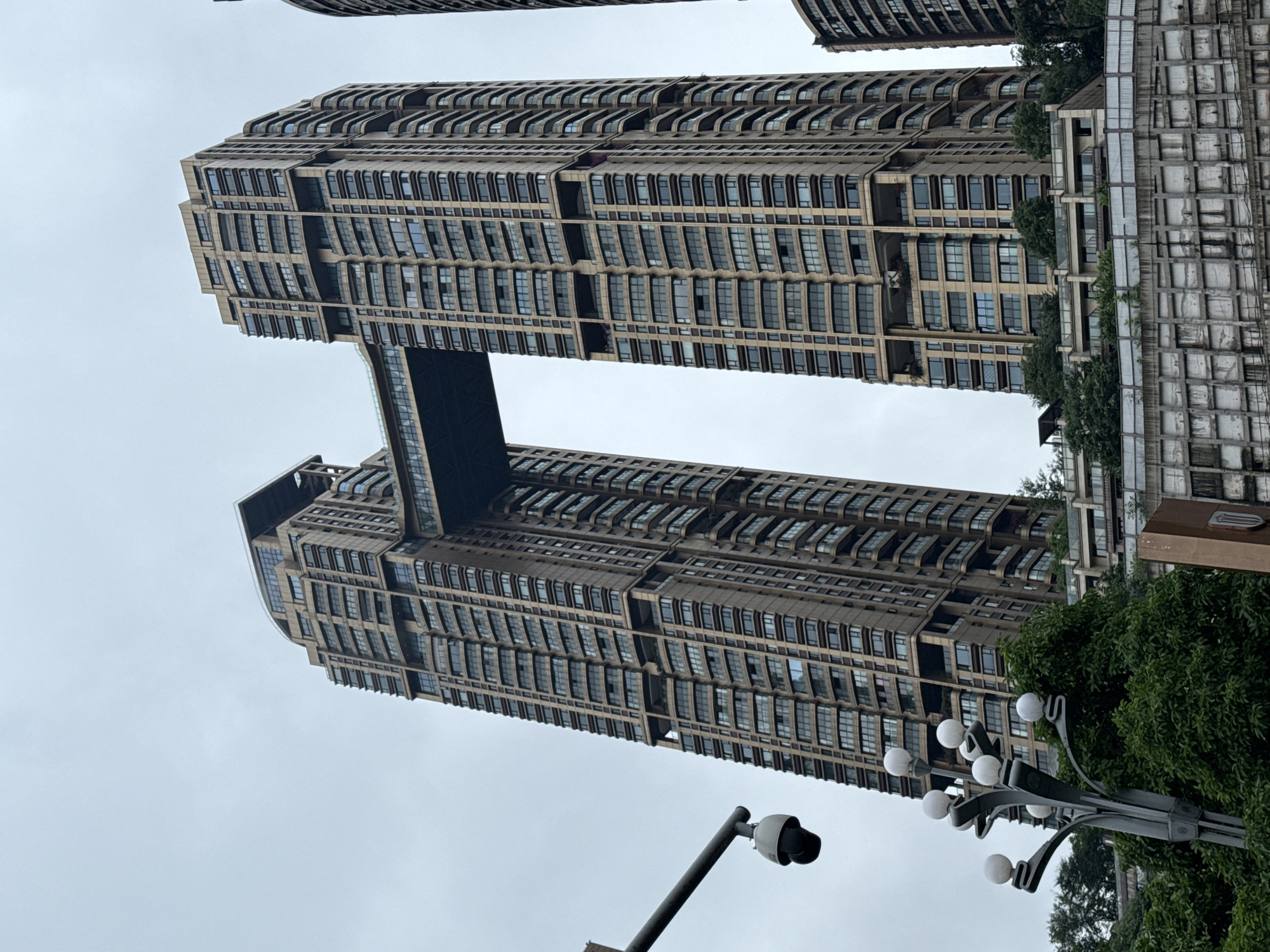 The image shows a low-angle view of two tall, brown buildings connected by a sky bridge. The buildings feature numerous windows and a tiered design. A street lamp with multiple spherical lights and a security camera stand in the foreground. Green trees are visible at the base of one building. The sky is overcast. The location is Nan'an District, China.