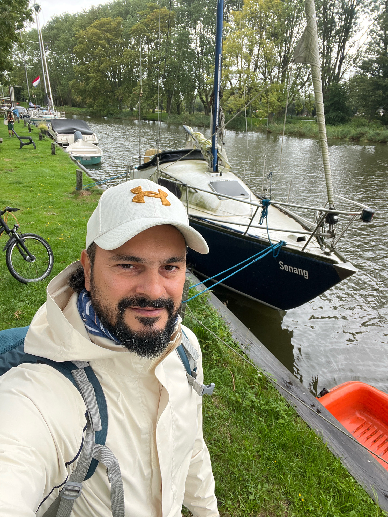 A man in a beige jacket and white baseball cap takes a selfie by the water in Edam, Netherlands. He has a beard and is wearing a backpack. Behind him, several boats are moored along the canal. One of the boats is a sailboat with the name "Senang" visible on its hull. The scene appears to be a marina or a canal-side park, with grass and trees lining the water's edge. The sky is overcast, suggesting a cloudy day.