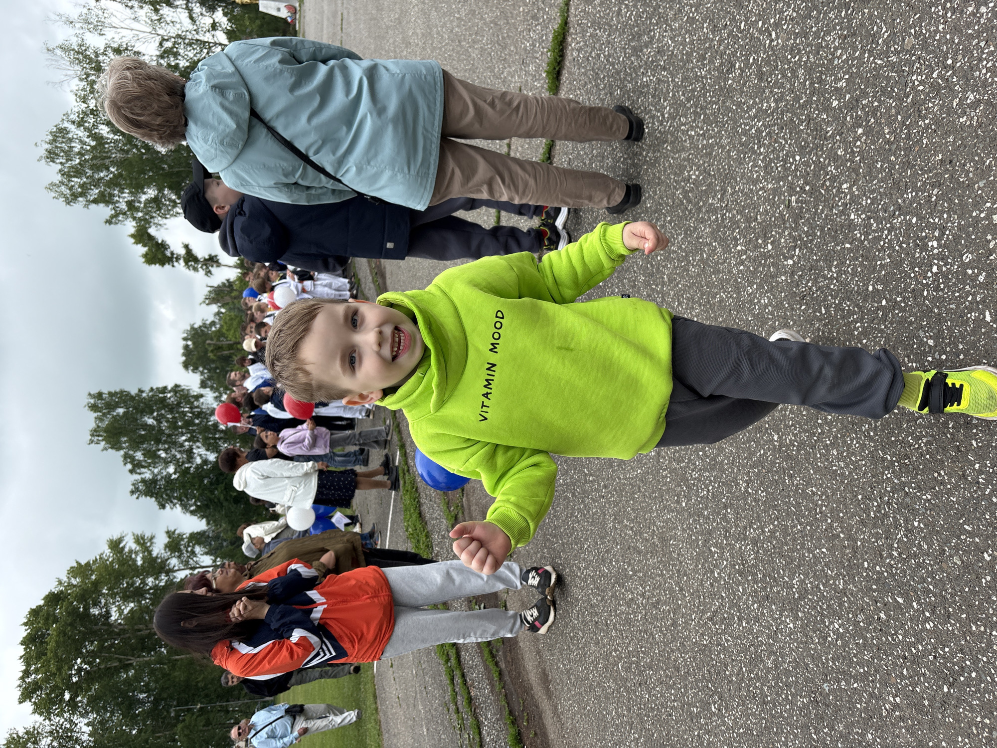 An outdoor scene in Kuyeda, Russia, features a young boy in the immediate foreground, centered and walking towards the camera. The boy, approximately 5-7 years old, has light brown hair, blue eyes, and an open-mouthed smile. He wears a bright lime-green hooded sweatshirt with "VITAMIN MOOD" printed in black, dark grey sweatpants, and black shoes with neon yellow accents and socks. His fists are loosely clenched.
Behind the boy, an assembly of individuals, primarily adults and children, occupies the midground and background. To the left, a woman with long dark hair, wearing an orange, white, and navy blue athletic jacket and light grey sweatpants, stands with clasped hands. Behind her, a man in a light blue shirt and sunglasses walks. To the right, the back of a woman in a light blue hooded jacket and khaki pants is visible, along with a boy in a black cap and dark jacket.
Further back, a crowd includes children dressed in white martial arts uniforms (gis), suggesting a sports event or demonstration. Red, white, and a partially visible blue balloon are interspersed among the attendees. The ground consists of dark asphalt with white aggregate, bordered by green grass and numerous trees in the background. An overcast sky is visible above. On the far right background, a stone monument with a golden element on top is discernible.