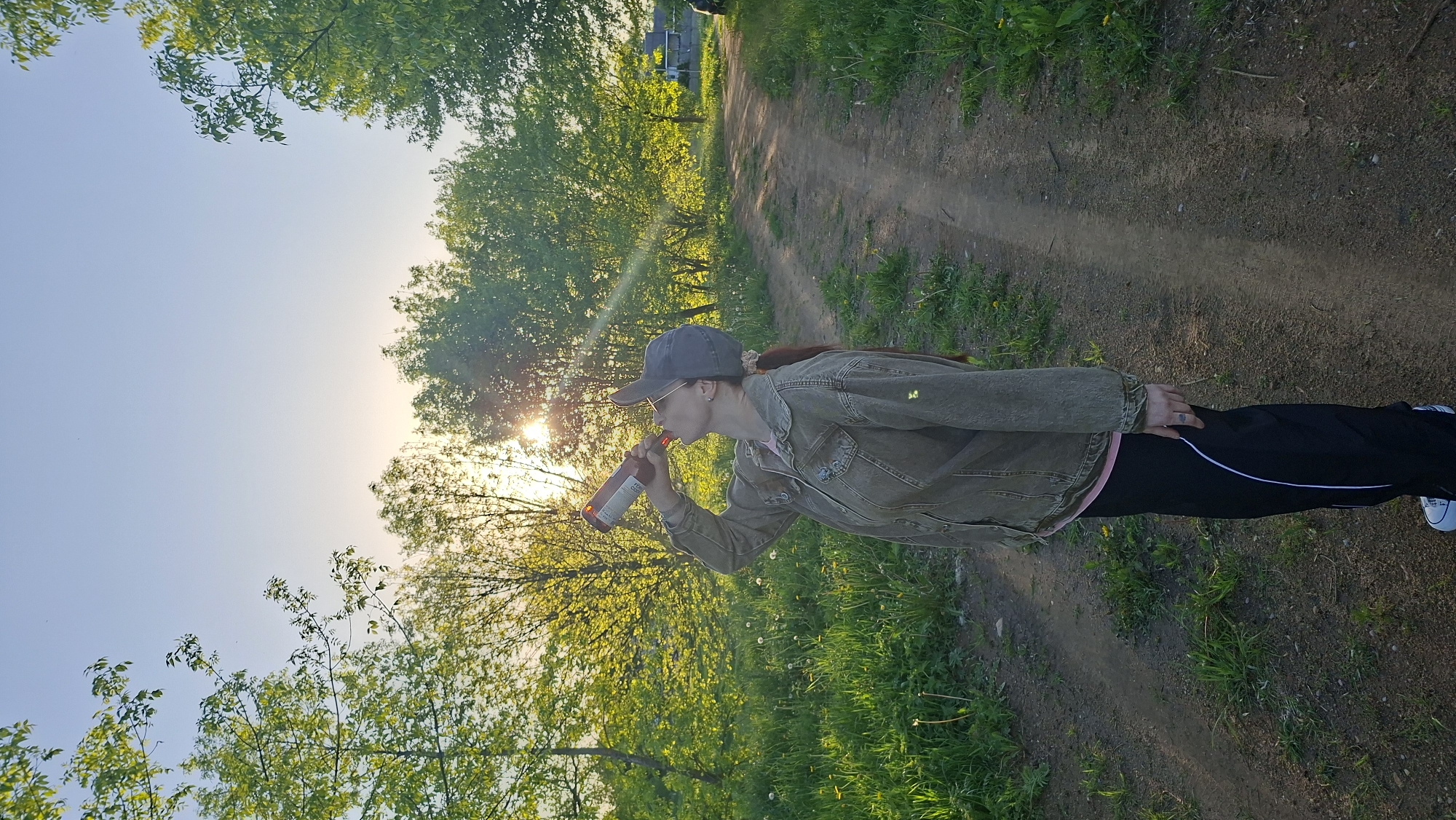 A person stands on a dirt path in Забалоцкі сельскі Савет, Belarus, drinking from a brown bottle. The individual is wearing a gray baseball cap, a tan jacket, and black pants with a white stripe. Green trees and foliage surround the path. The sun shines brightly in the sky.