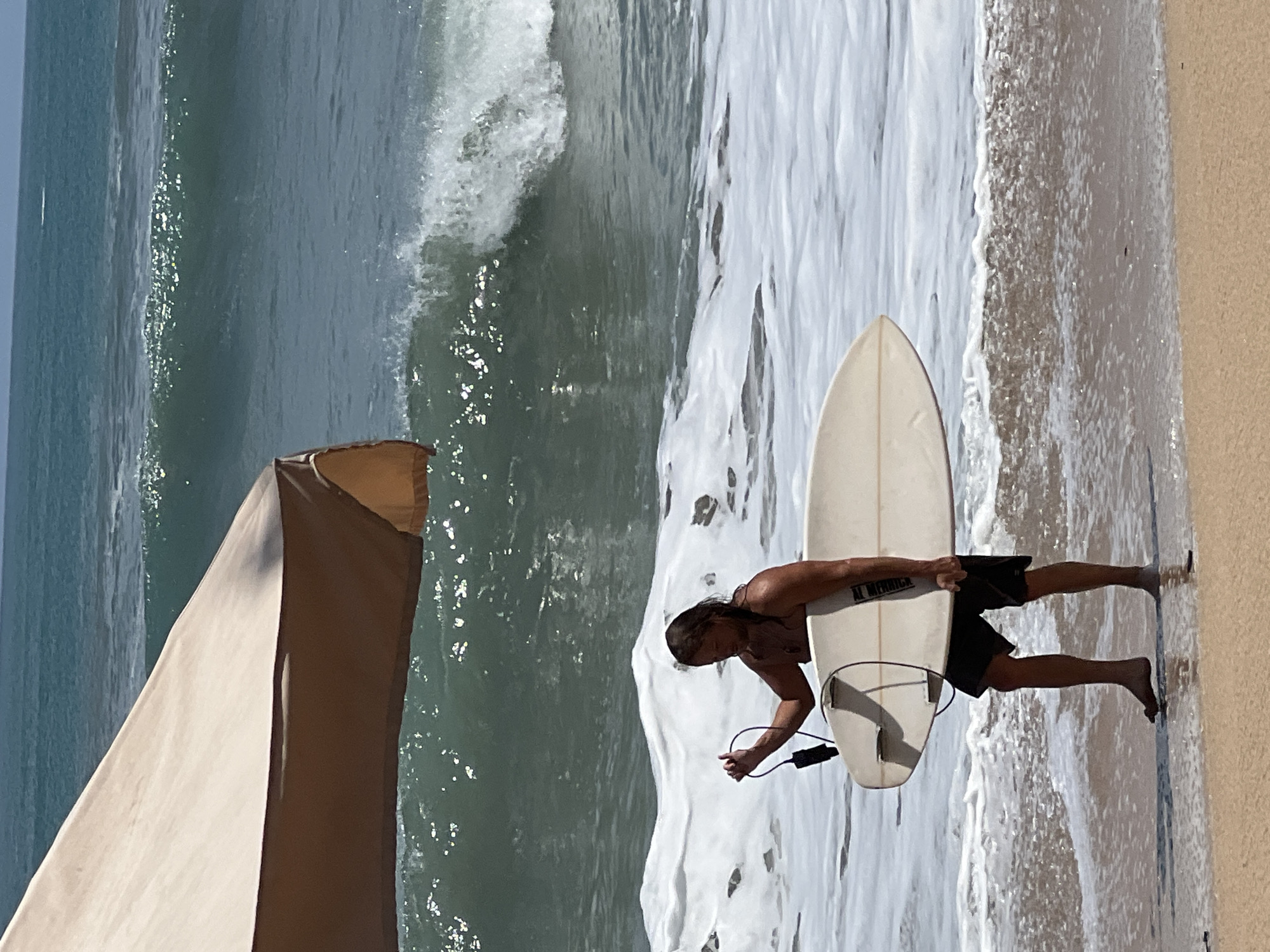 A surfer, wearing black shorts, carries a white surfboard and leashes into the shallow water of the ocean. The surfer has long, light brown hair and a tattoo on their right arm. The surfer is standing in the surf zone with waves breaking around them, creating white foam. In the background, a large wave is cresting, and the ocean stretches out to the horizon under a clear sky. A light-colored umbrella is partially visible in the upper left corner of the image. The scene appears to be the beach in Kuta, Indonesia, known for its surfable waves and sandy shores.