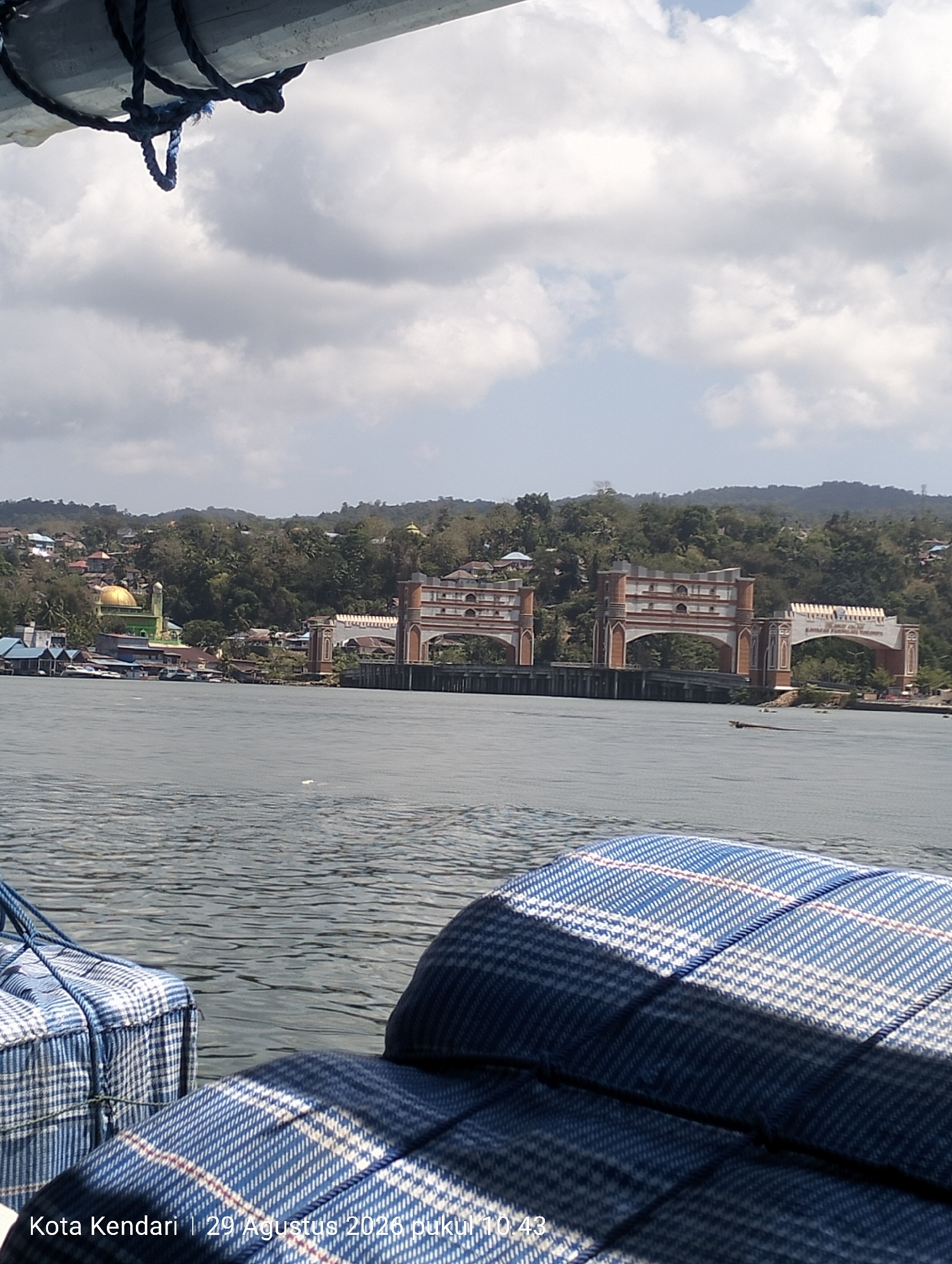 The image shows a view from a boat on the water, looking towards a city on the coast. In the foreground, there are two blue and white checkered cushions. The water is calm with ripples. In the middle ground, there is a body of water with some small debris visible. On the far bank, there is a cityscape with buildings and lush green hills in the background. A prominent feature is a large, arched bridge or gateway structure with ornate brickwork. To the left of the bridge, a green mosque with a golden dome is visible. The sky is partly cloudy with bright sunlight. The lower left corner of the image contains text that reads "Kota Kendari | 29 Agustus 2026 pukul 10:43", indicating the location as Kendari, Indonesia, and the date and time.