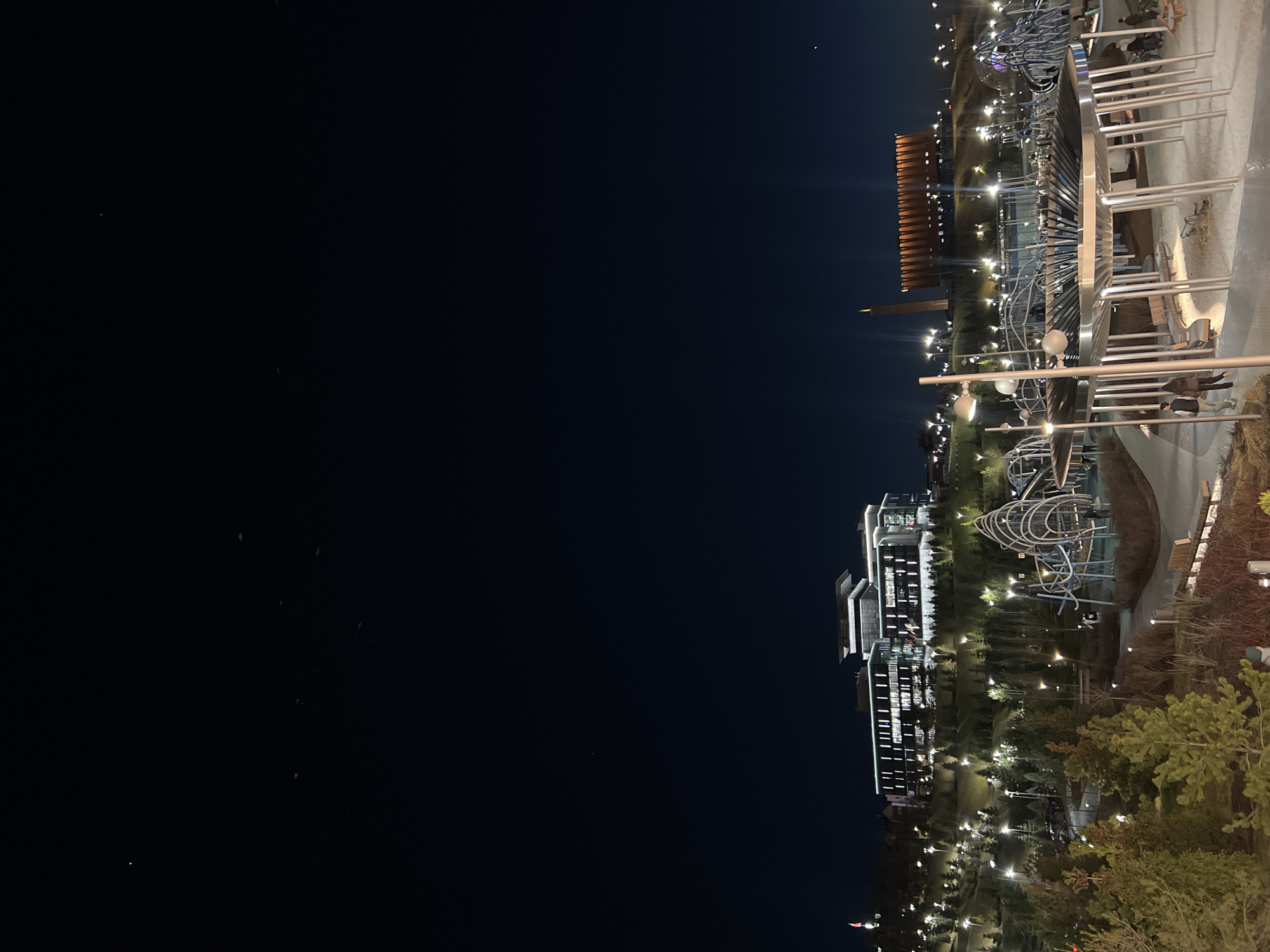 At night, the image captures the cityscape of Kazan, Russia. The left side of the image is predominantly dark, presumably a body of water. On the right side, lights illuminate several buildings, trees, and an open space featuring modern metallic art installations. The architecture visible includes a multi-storied building with lighted windows and a taller tower structure in the distance. The foreground shows vegetation alongside the walking paths. People can be observed walking on the paths near the art pieces, under the lights.
