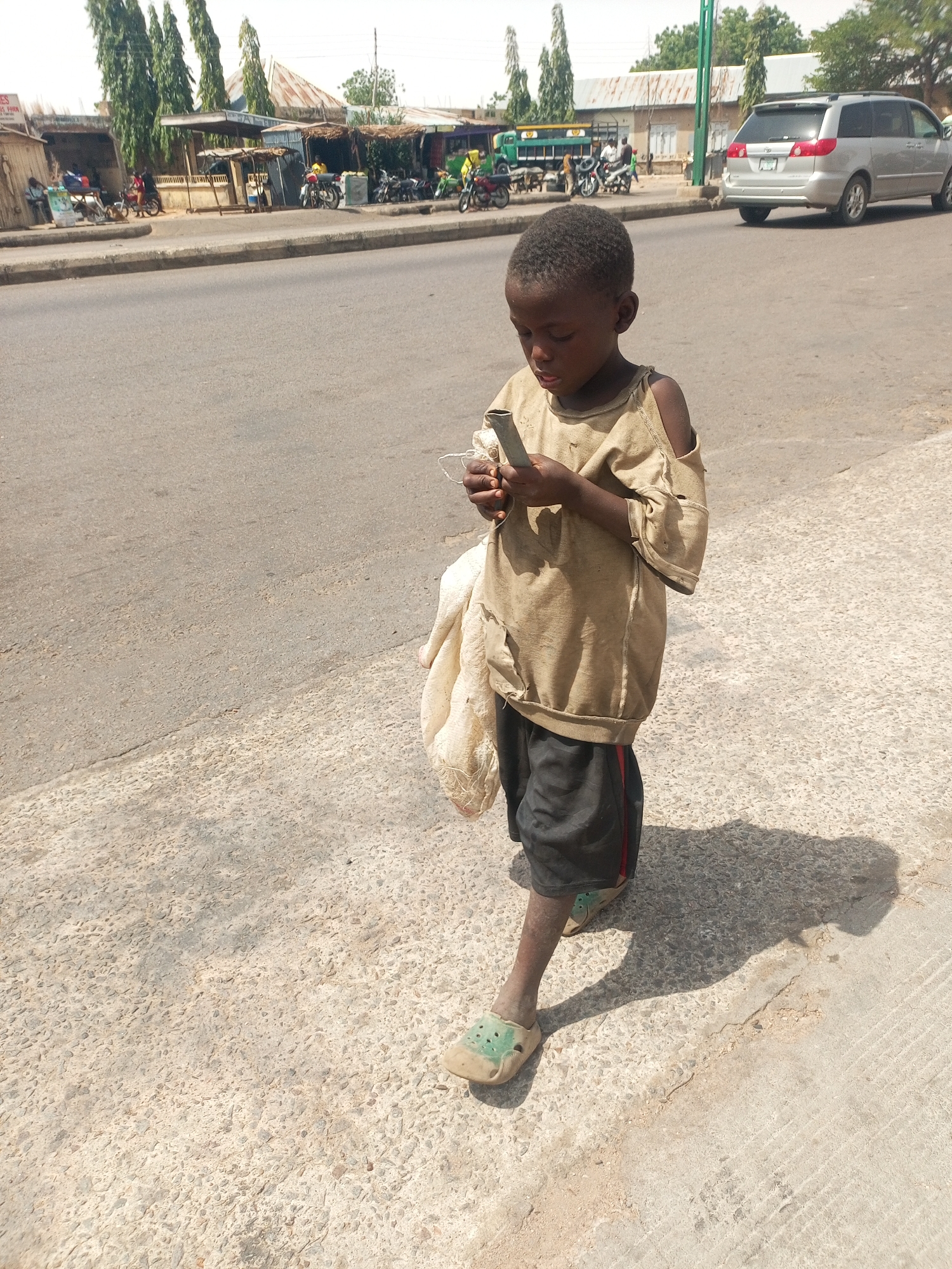 A young, dark-skinned boy stands on a concrete sidewalk in Gombe, Nigeria. He wears a tattered tan shirt with ripped shoulders, black shorts with a red stripe, and worn green Crocs. He holds a small, rectangular object and looks down at it. A dirty white cloth bag hangs from his left arm. Behind him is a paved road with vehicles, including a silver minivan, motorcycles, and a green truck. Buildings and trees line the street. The lighting suggests it is daytime.